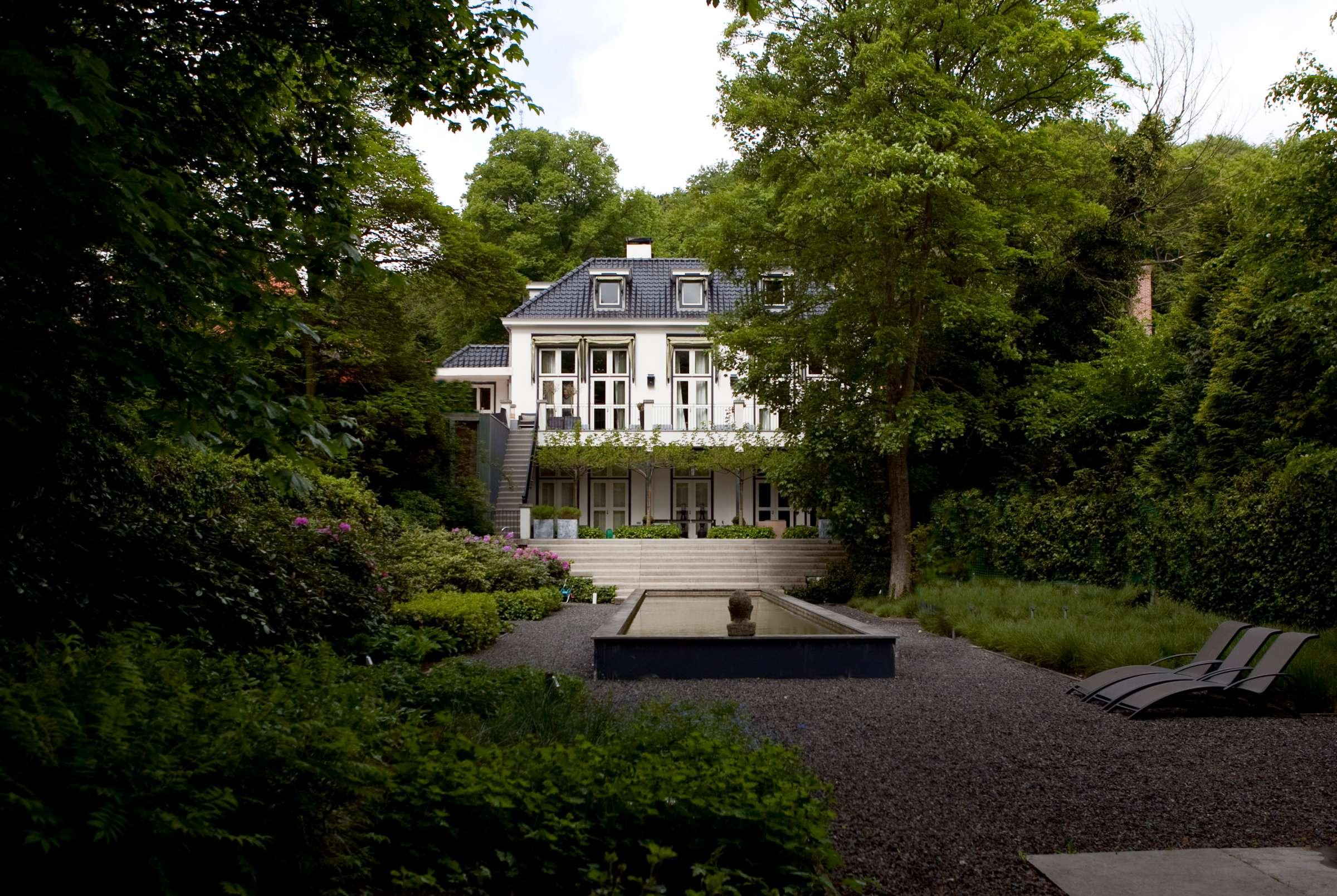 Villa omgeven door bomen en een tuin, met een trap en een waterpartij vooraan.