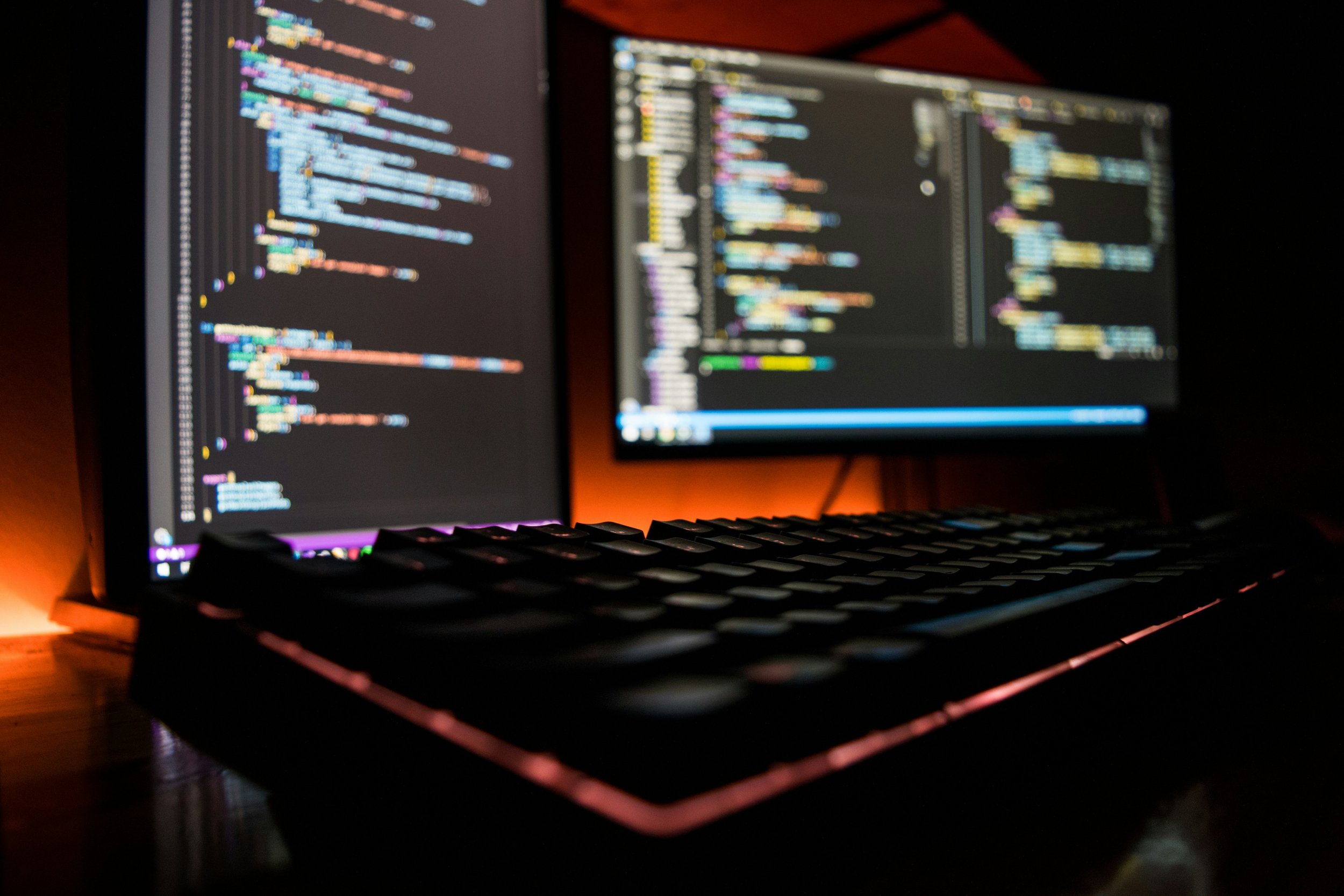 Close-up of a dual computer monitor setup displaying colorful code editing or programming environment in a dimly lit room.