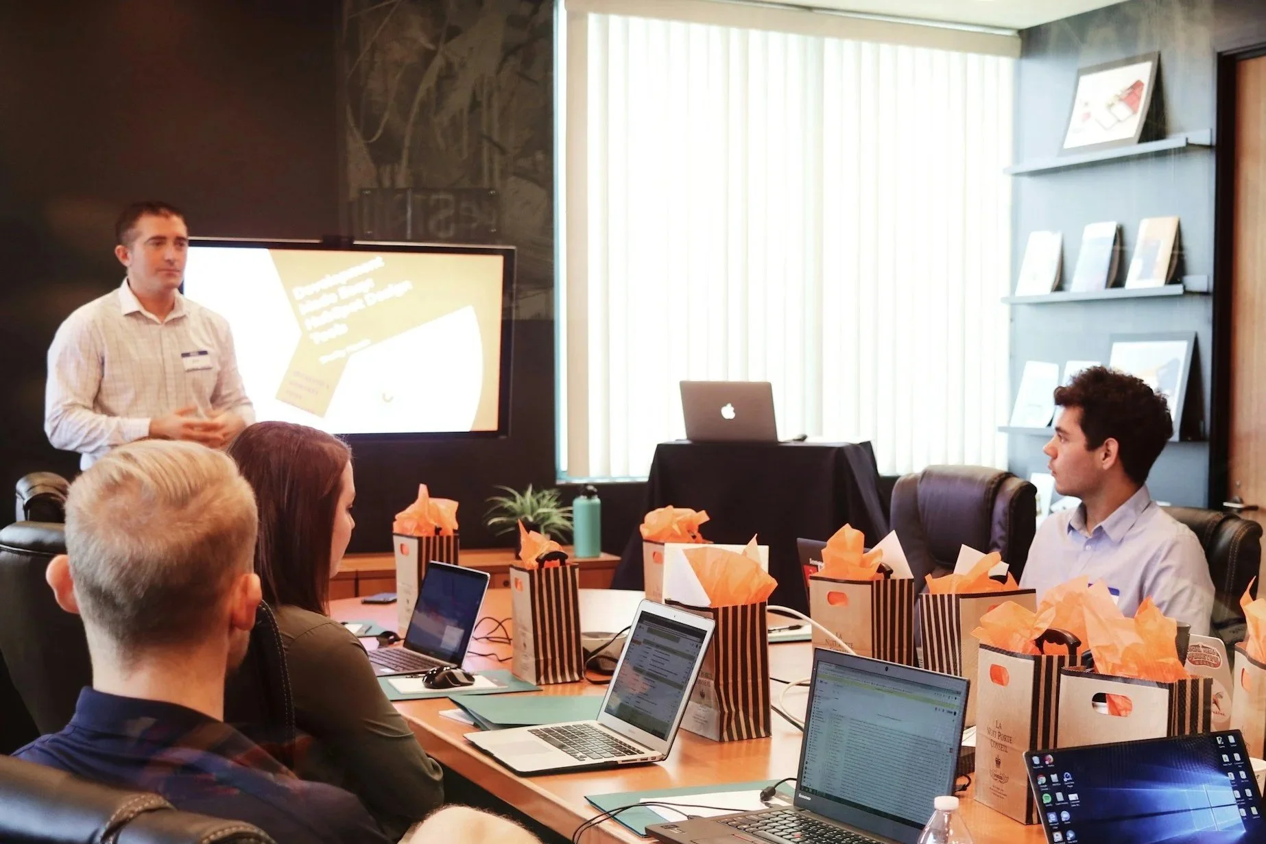 A business meeting in a conference room with a man standing and presenting in front of a screen, while seated attendees work on laptops and observe the presentation, with gift bags on the table.