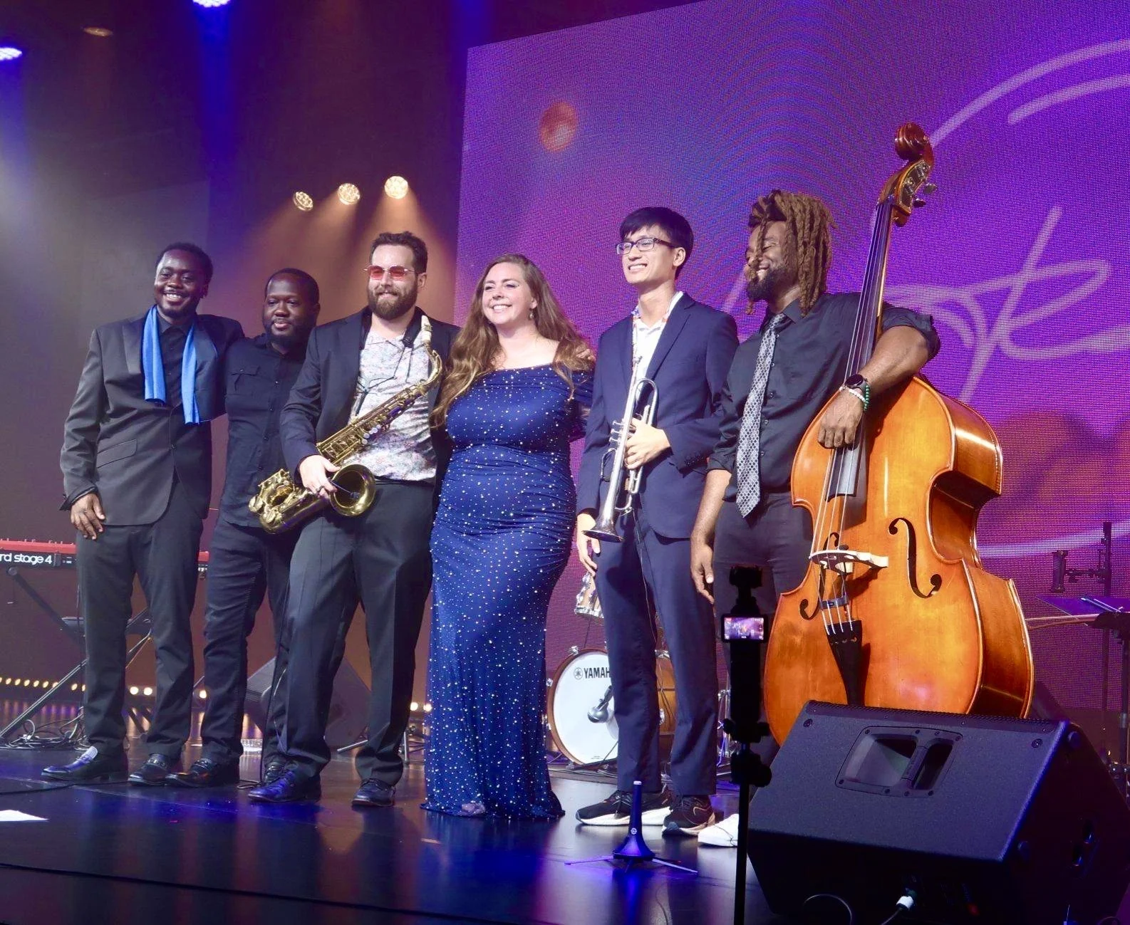 Six musicians standing on stage after a performance, smiling and posing for a photo. They are dressed in formal and stylish attire, holding their musical instruments, including a saxophone, trumpet, and double bass, with a backdrop of stage lighting and a large screen.