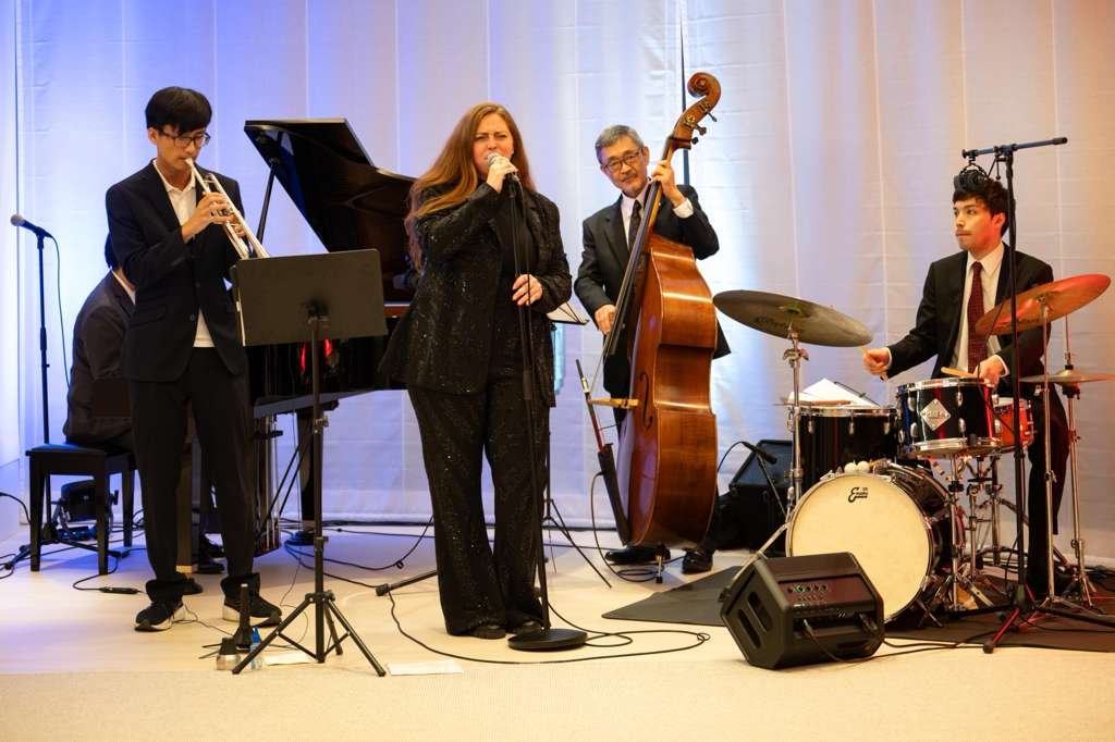 A jazz band performing on stage, featuring a woman singing into a microphone, a double bass player, a drummer, a pianist, and a trumpet player.