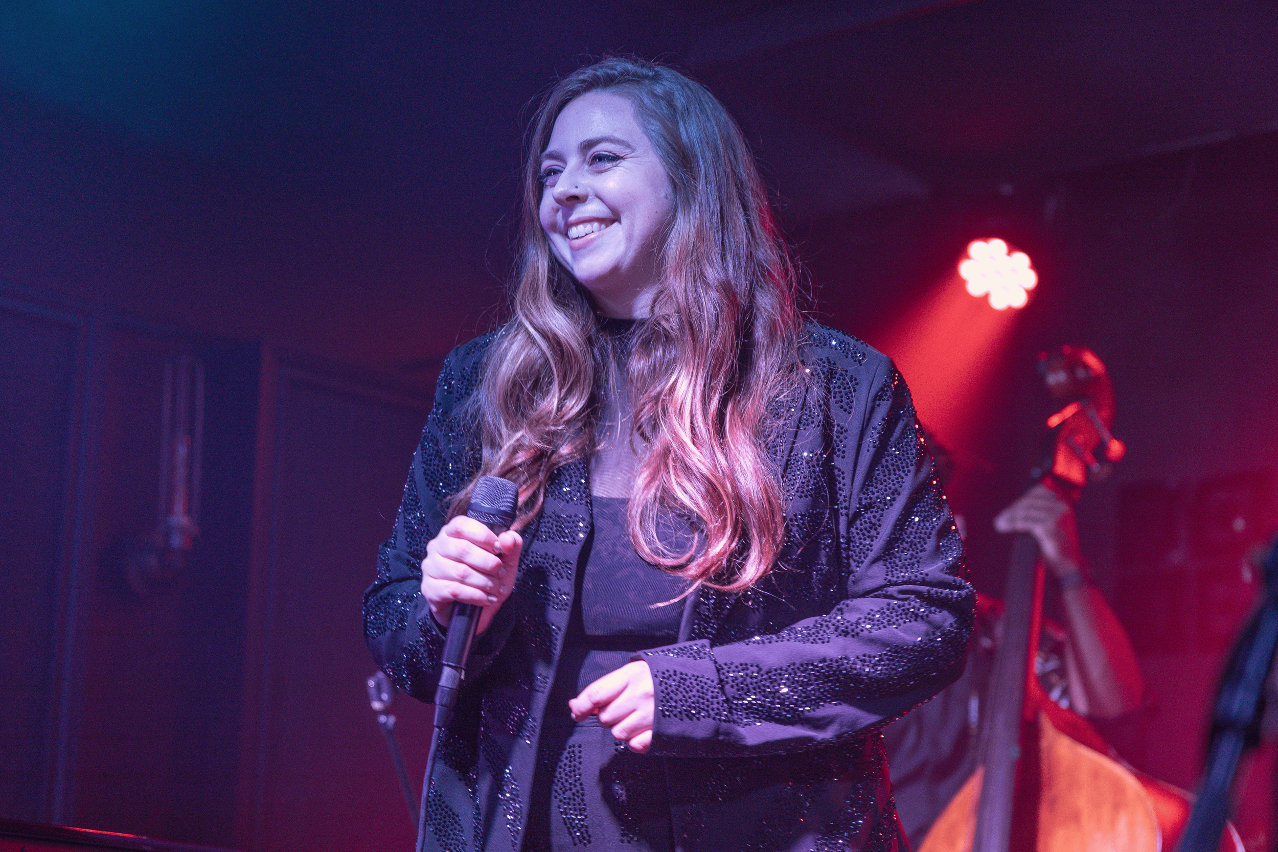 A woman with long, wavy hair holding a microphone on stage. She is smiling and wearing a black, sequined jacket. There are colorful stage lights in the background.