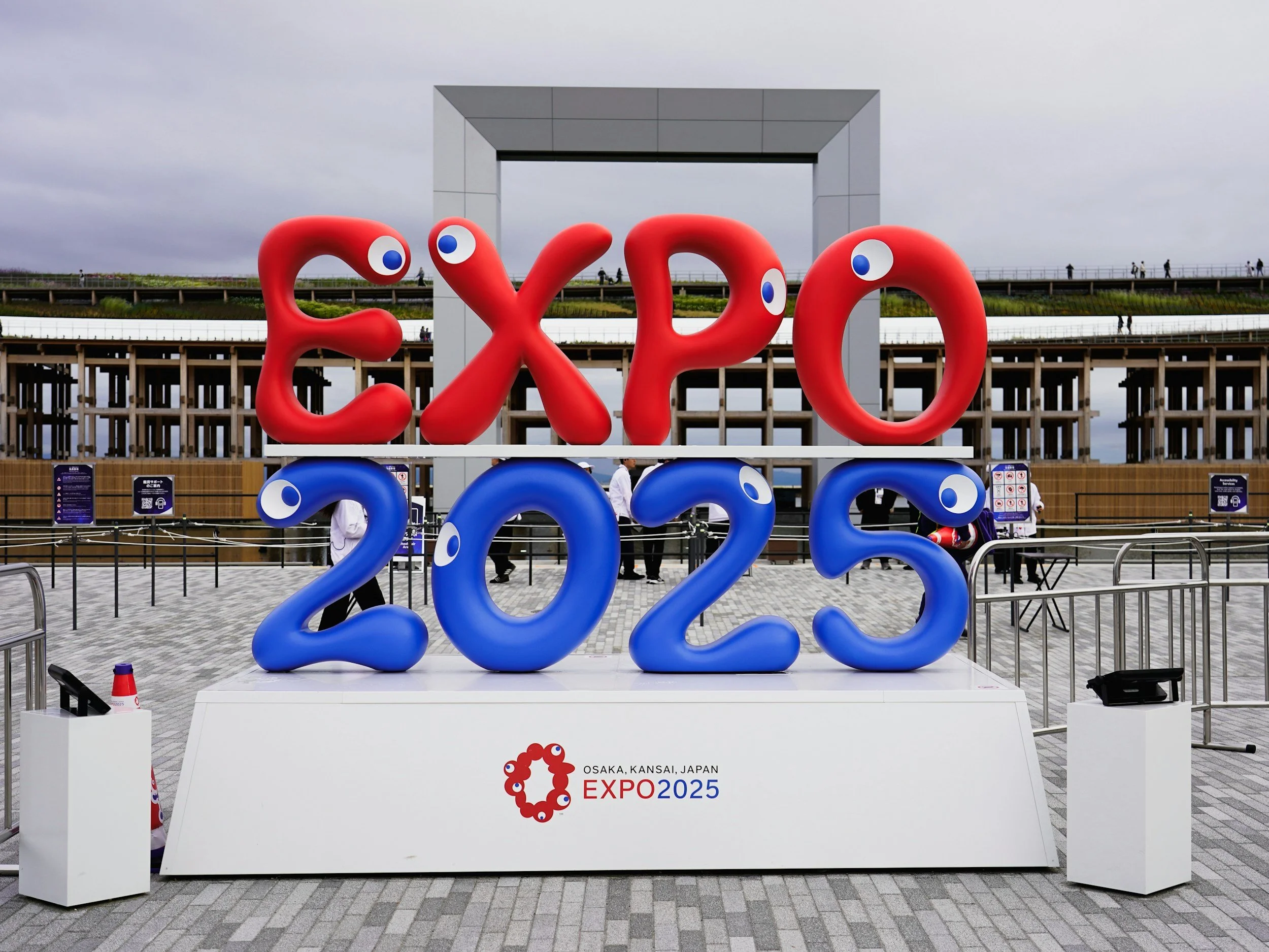 Large red and blue cartoonish letters spelling 'EXPO' with eyes, and the year '2025' in blue below, on a white platform with a sign indicating Osaka, Kansai, Japan, and a logo with a red circle of connected dots. Behind the display, there is an outdoor setting with a modern structure, walking people, and a cloudy sky.