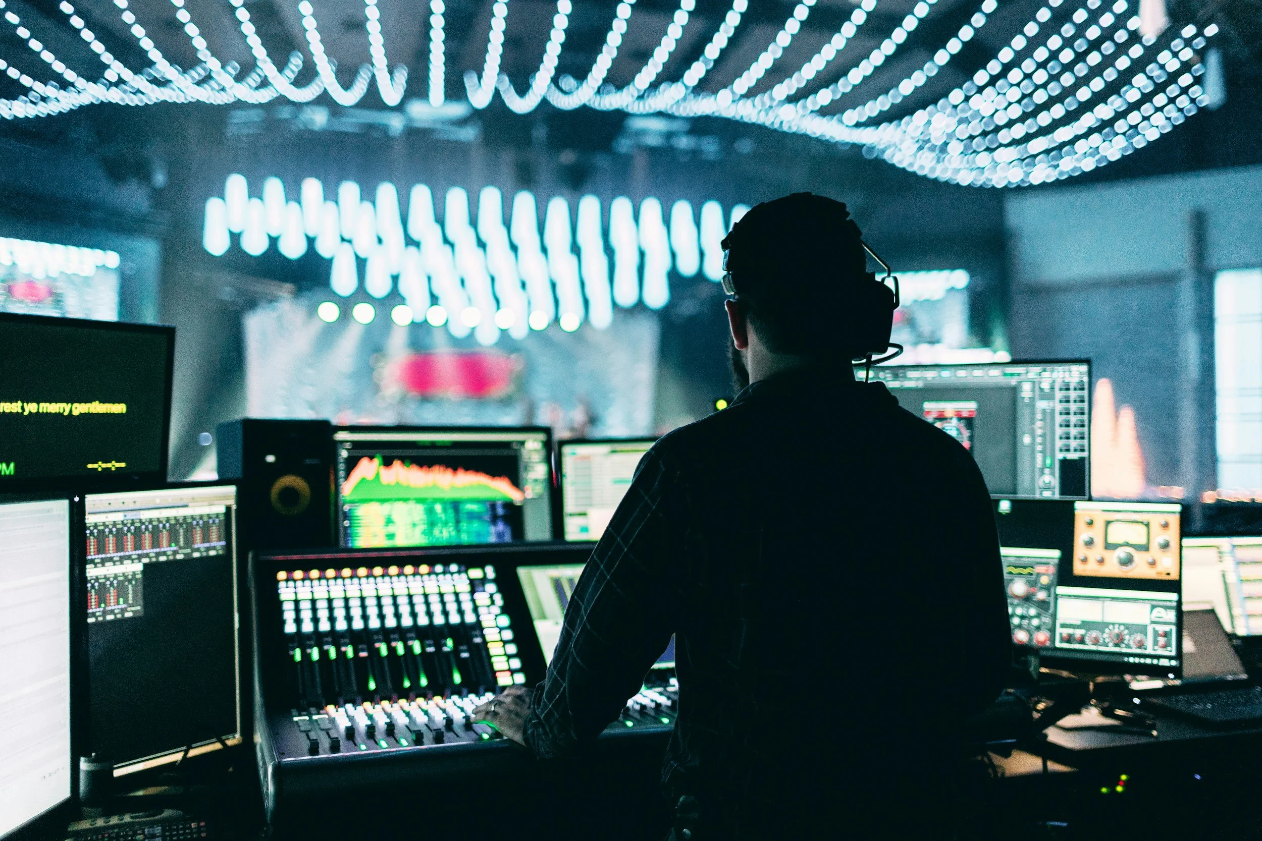 A person working in a control room with multiple computer screens displaying colorful graphs and data, illuminated by bright blue and white LED lights on the ceiling.