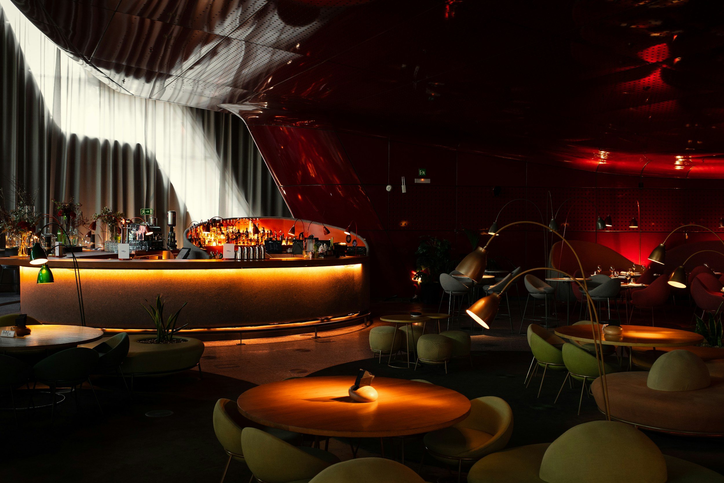 A dimly lit modern lounge or bar area with a curved bar counter, various bottles of alcohol, plants, and comfortable seating, with a dark red and black color scheme and stylish lighting.