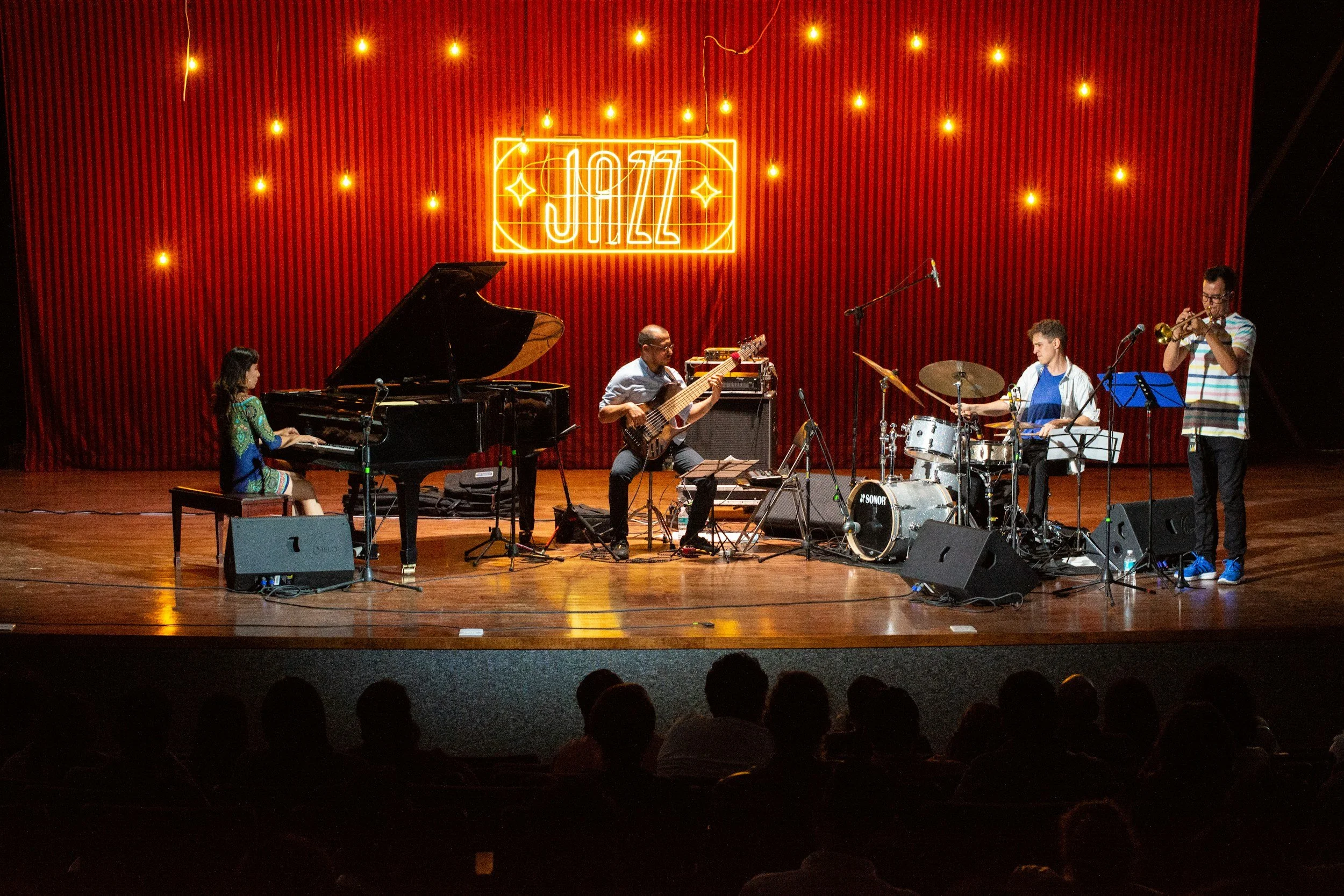 Jazz band performing on stage with a piano, bass guitar, drums, and trumpet in front of a red curtain with string lights and a neon sign that says "JAZZ."