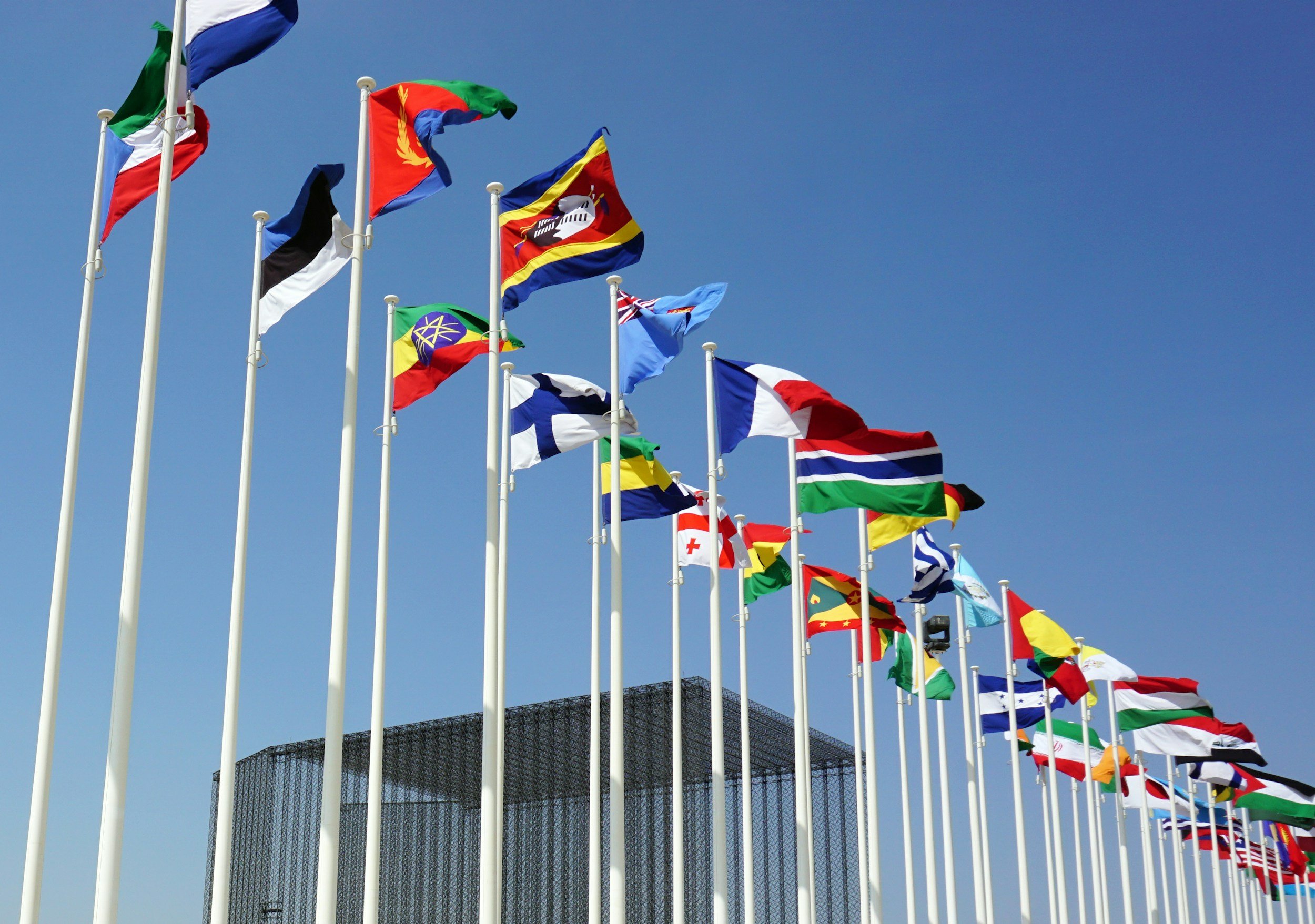 Multiple international flags on flagpoles flying against a blue sky, with a building in the background.