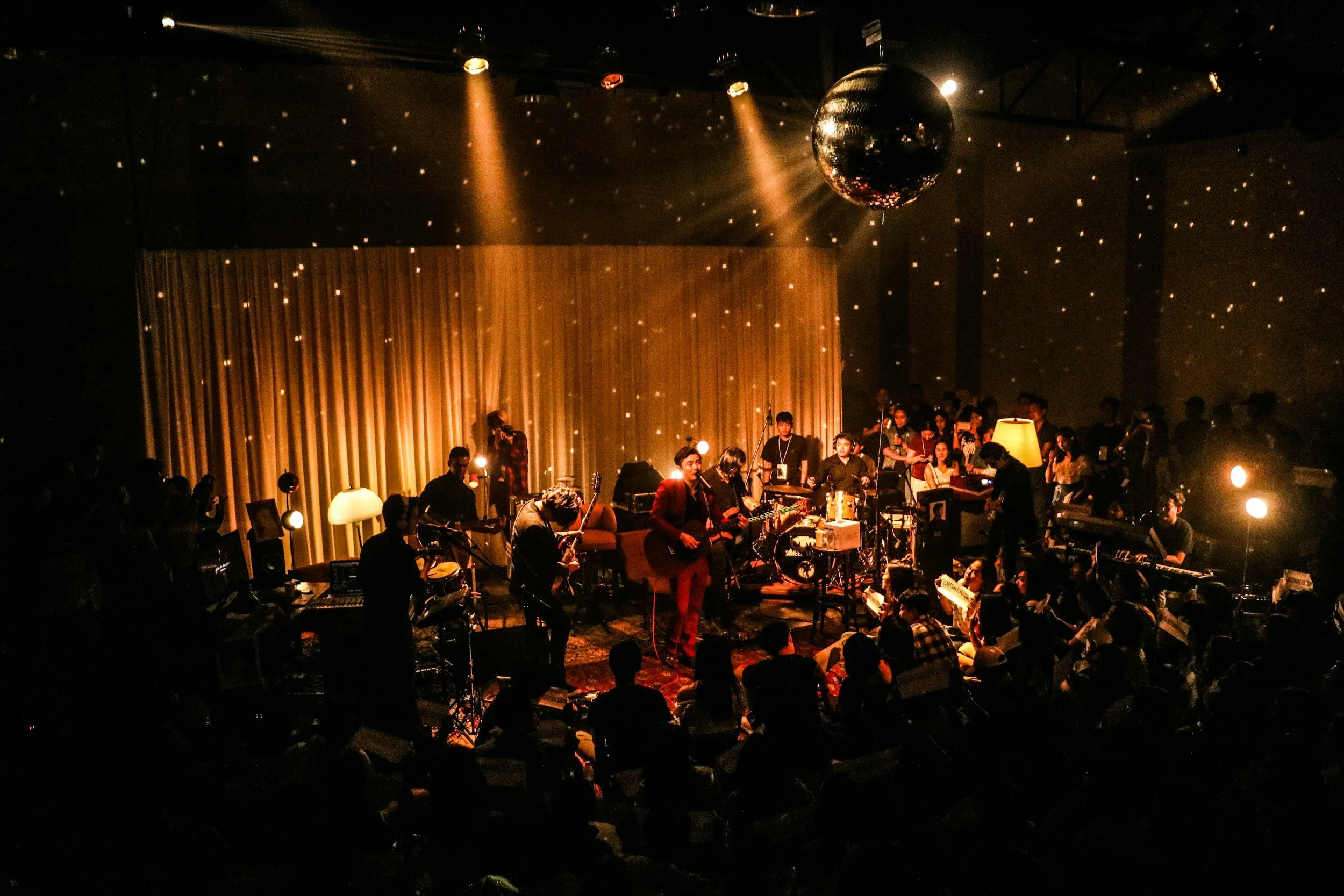 A dimly lit indoor concert featuring a live band performing on stage with a disco ball hanging from the ceiling, surrounded by an audience seated and standing around the stage.