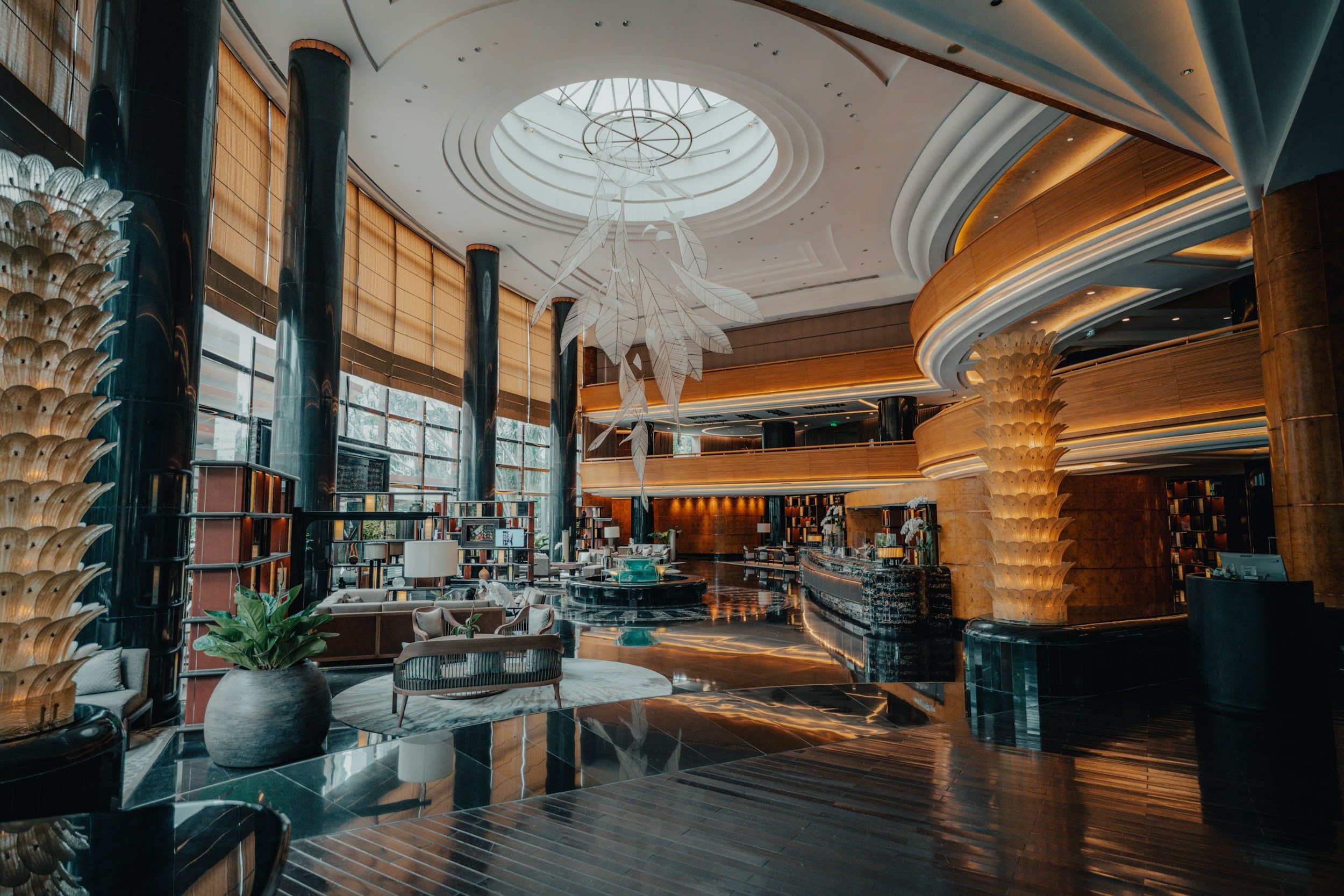 Interior of a luxurious hotel lobby with high ceilings, large windows, and modern decor. There is a large chandelier made of metallic leaves hanging from the ceiling, surrounded by tiered, curved balconies with warm lighting. The floor is shiny black marble with intricate patterns, and stylish seating arrangements are visible.