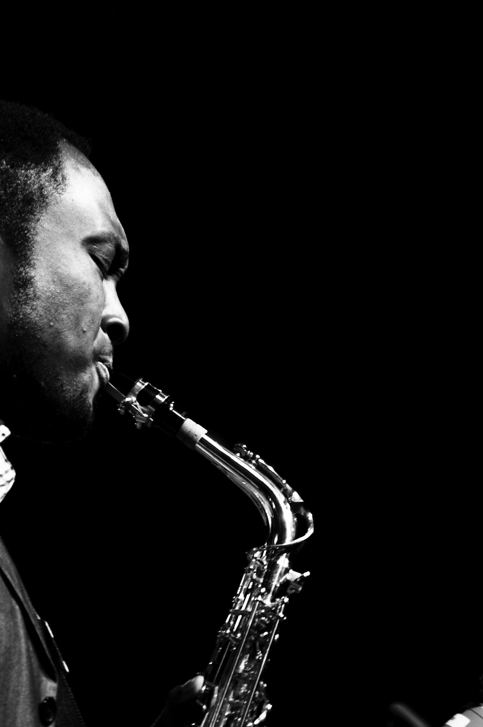 Close-up black and white photo of a person playing a saxophone, showing the person's face in profile with closed eyes and the saxophone in focus against a dark background.