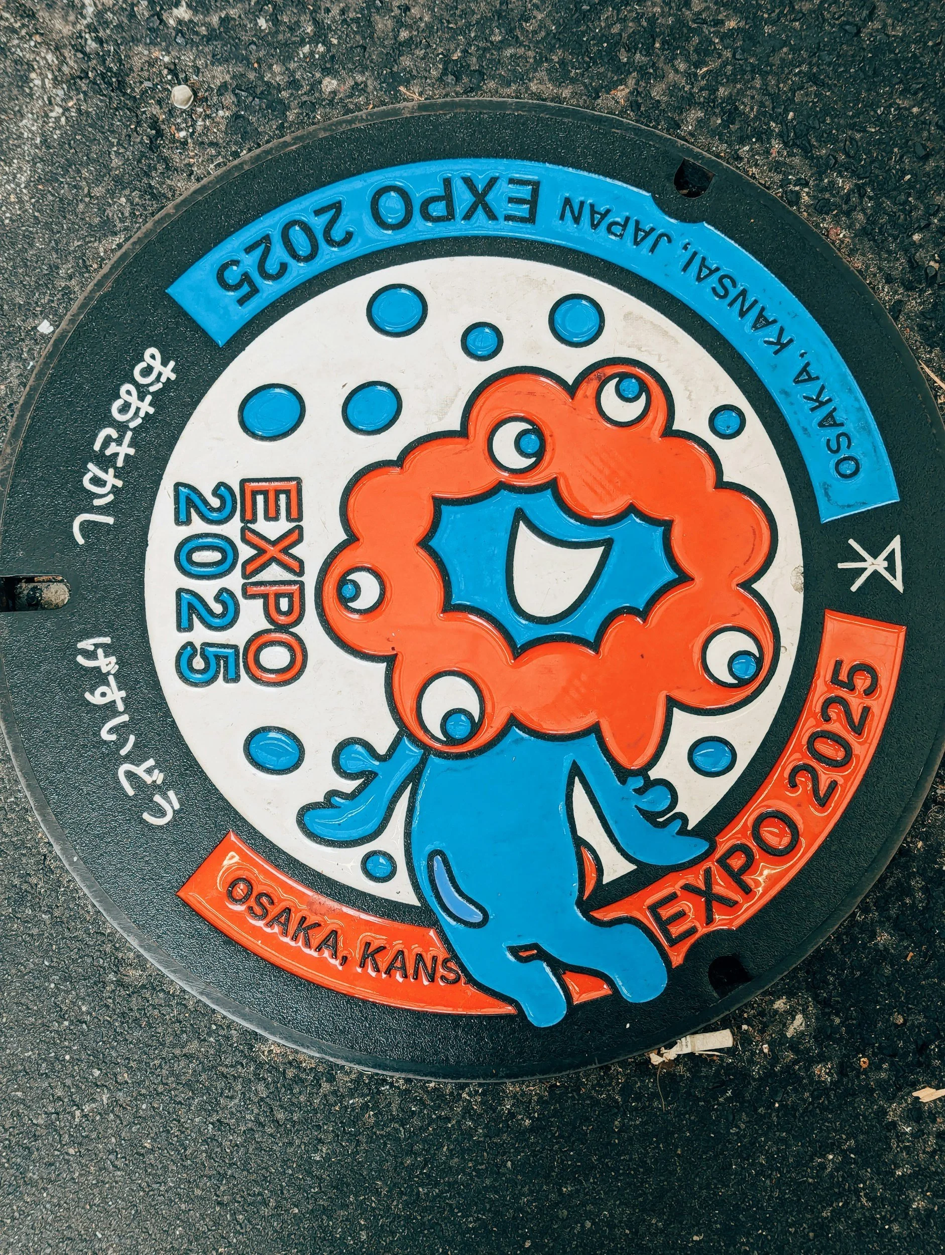 Colorful manhole cover with a smiling face and a floral design, located on a paved street surface in Osaka, Kansas, with engraved details and a date of 2025.