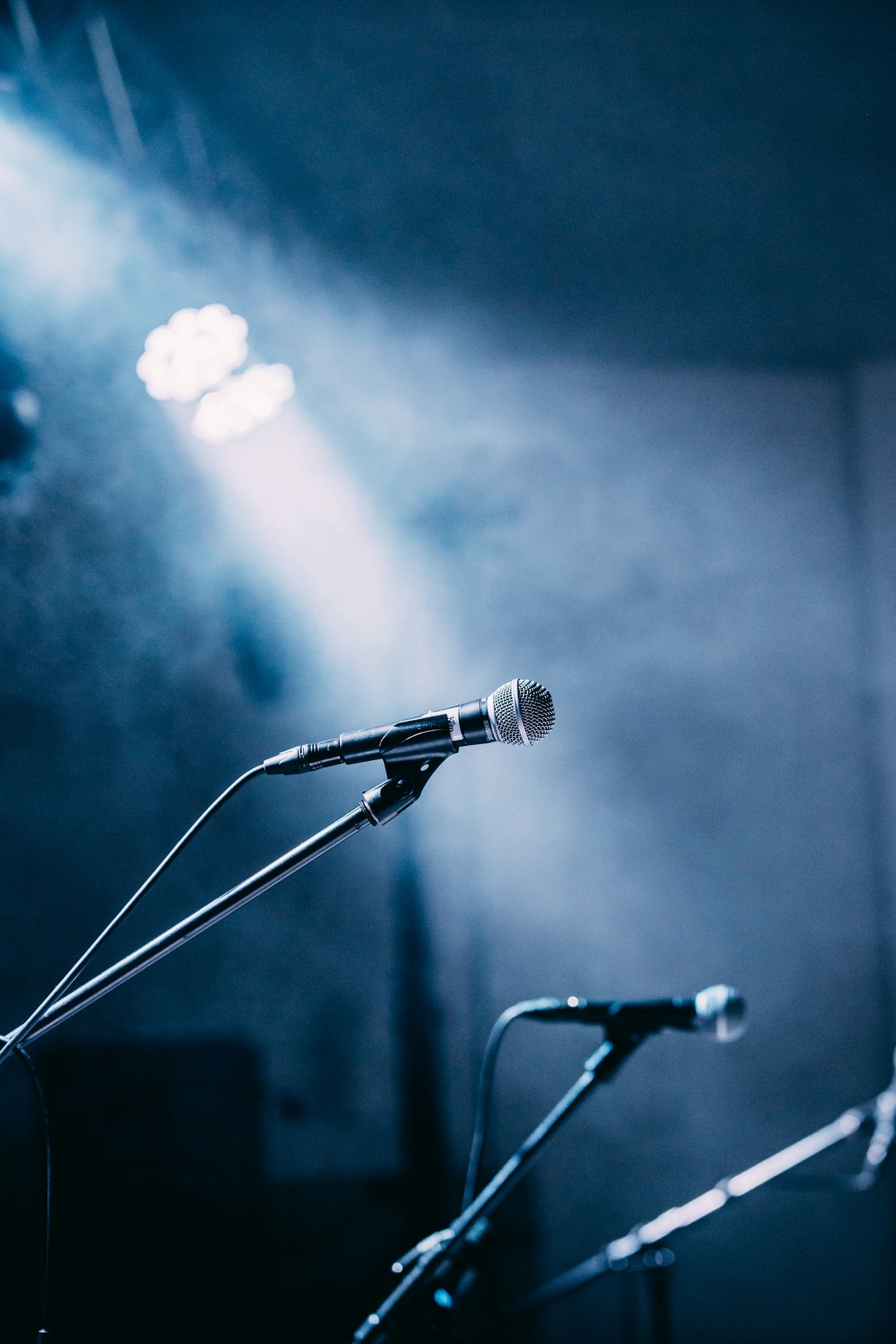 Close-up of multiple microphones on stands set up on a stage with blue lighting.