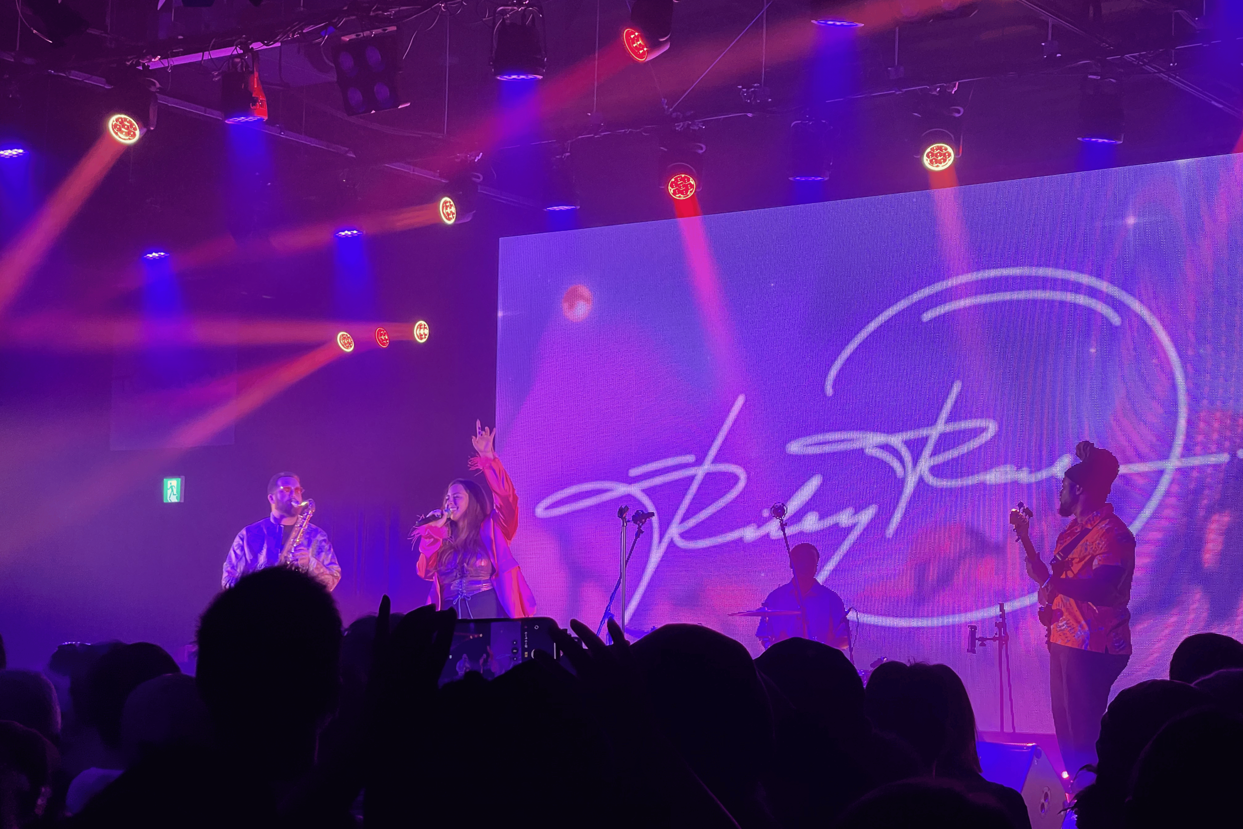 A live musical performance on stage with purple and red lighting. Two singers, a man playing the saxophone and a woman singing into a microphone are visible. Large screen in the background displays white cursive text.