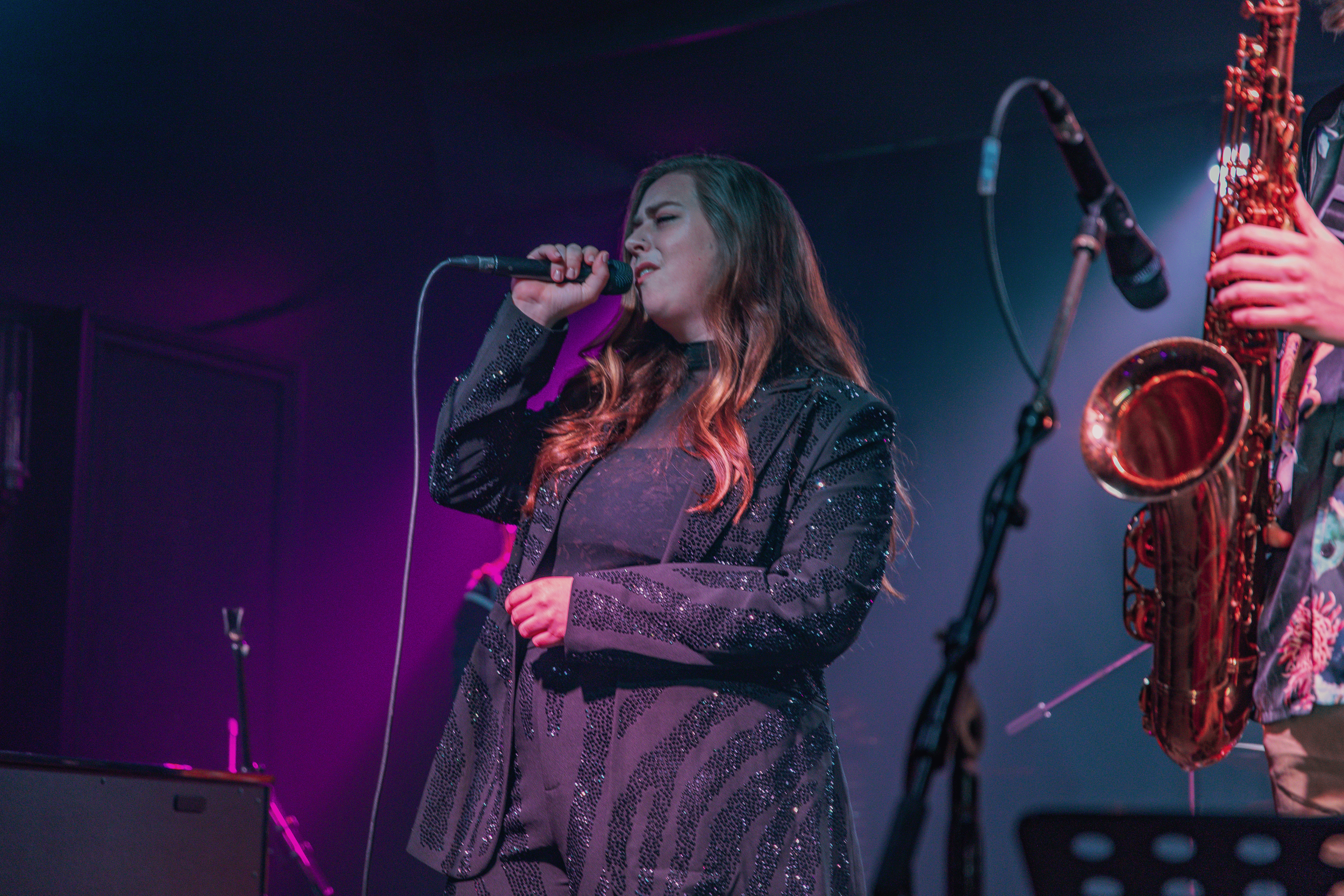 A female singer with long, wavy hair singing into a microphone on stage, wearing a black sequined jacket, with a saxophone player in the background.