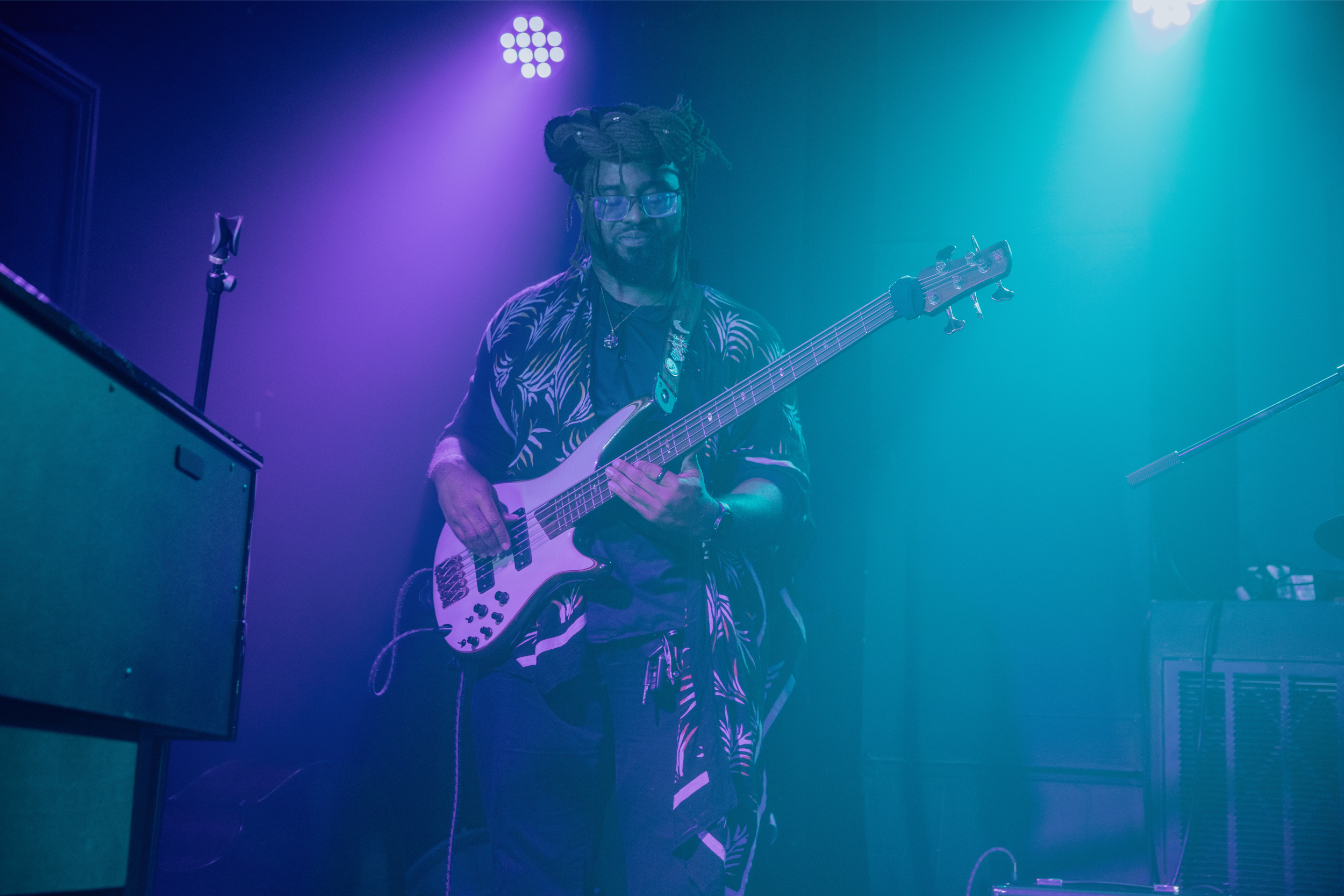 A musician with dreadlocks and glasses playing an electric bass guitar on stage with colorful purple and blue lighting.