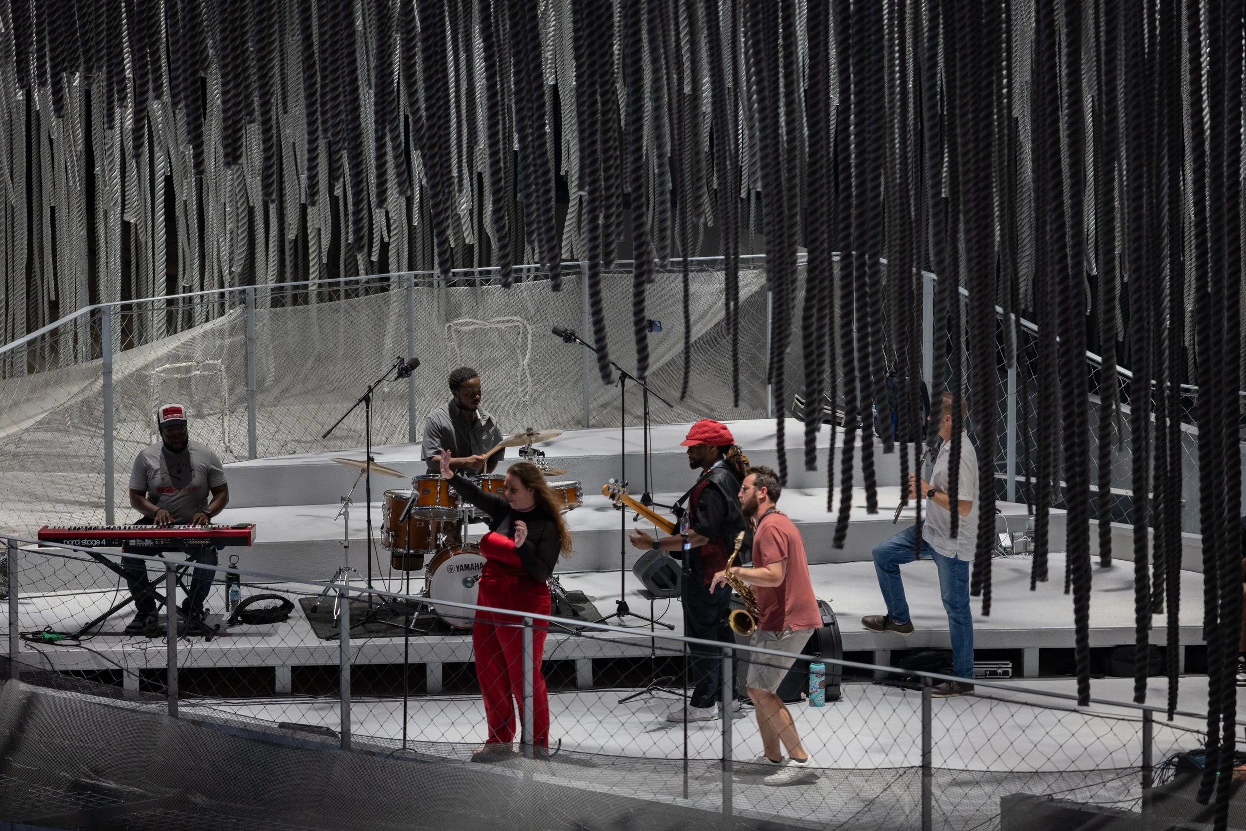 A music band performing on stage with various instruments, people singing and playing, seen through a mesh barrier.