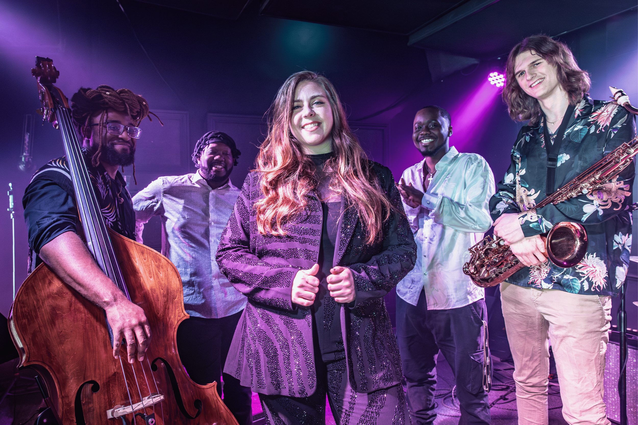 Six musicians standing on stage after a performance, with a purple backdrop and stage lights.