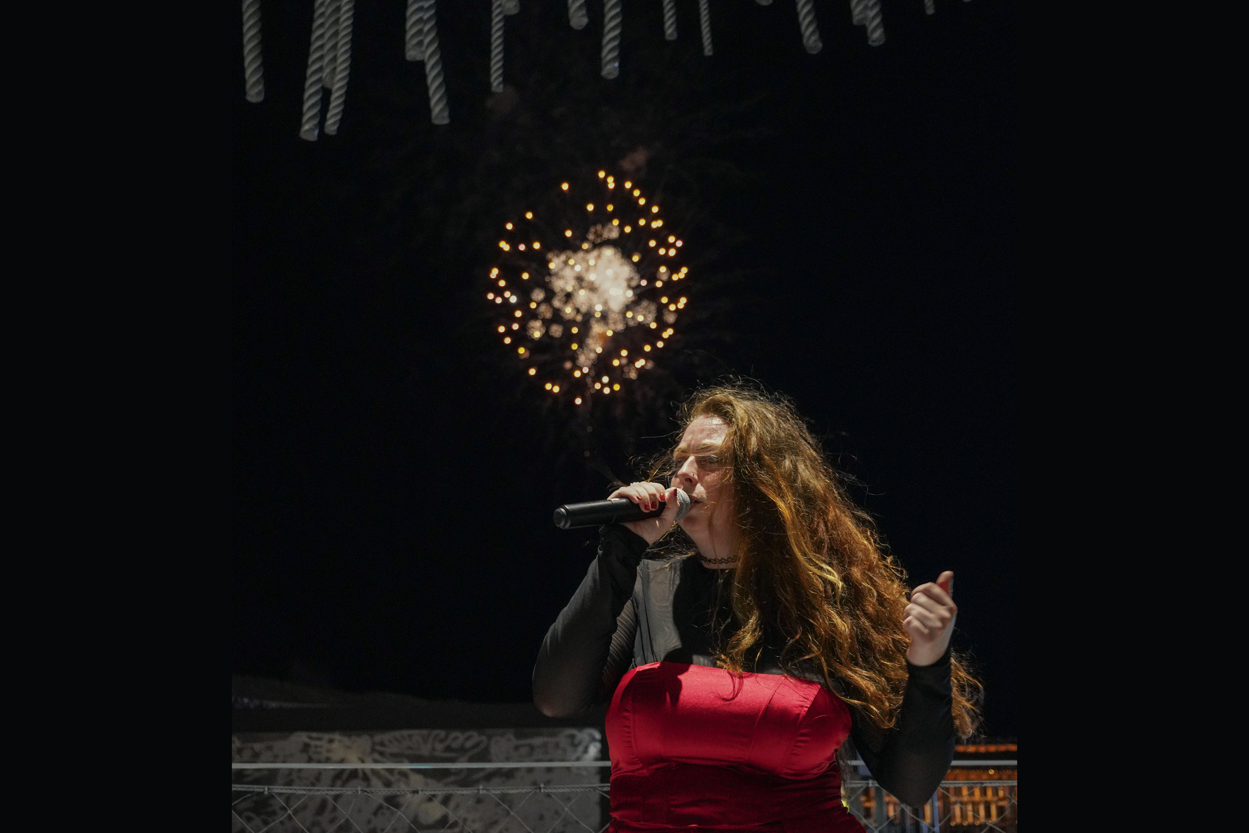 A woman with long curly hair singing into a microphone on stage, with fireworks in the dark sky behind her.