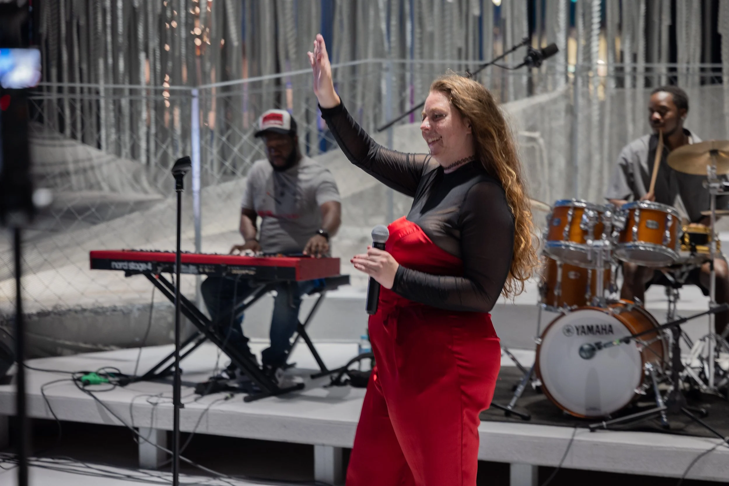A woman with long curly hair, dressed in a red and black outfit, holding a microphone, appears to be singing or speaking. Behind her, a band setup includes a keyboard, drums, and a music stand, with a drummer and a person at a keyboard playing on a s