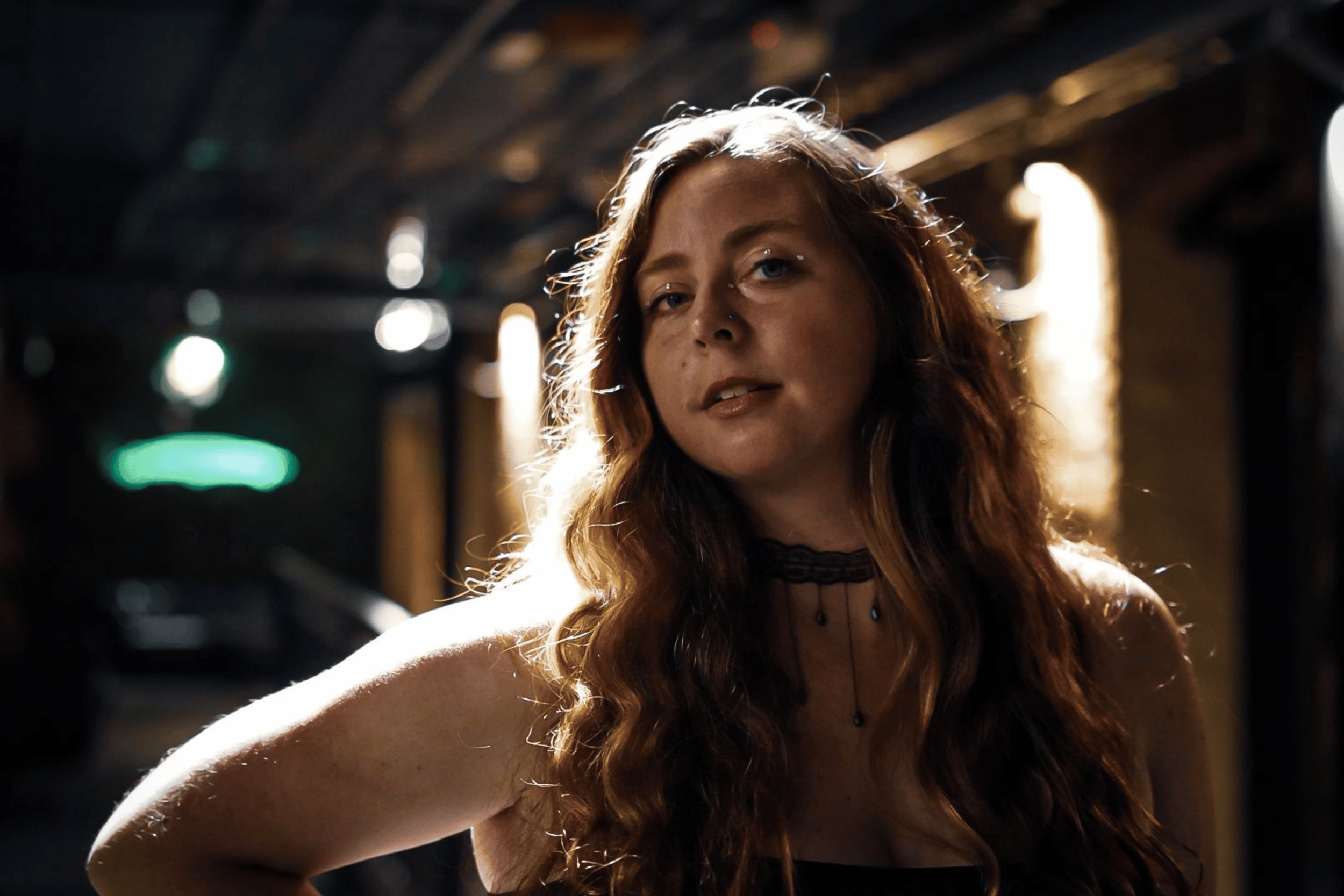 A woman with long wavy red hair and blue eyes, wearing a black choker and black top, standing in a dimly lit environment with blurry lights in the background.