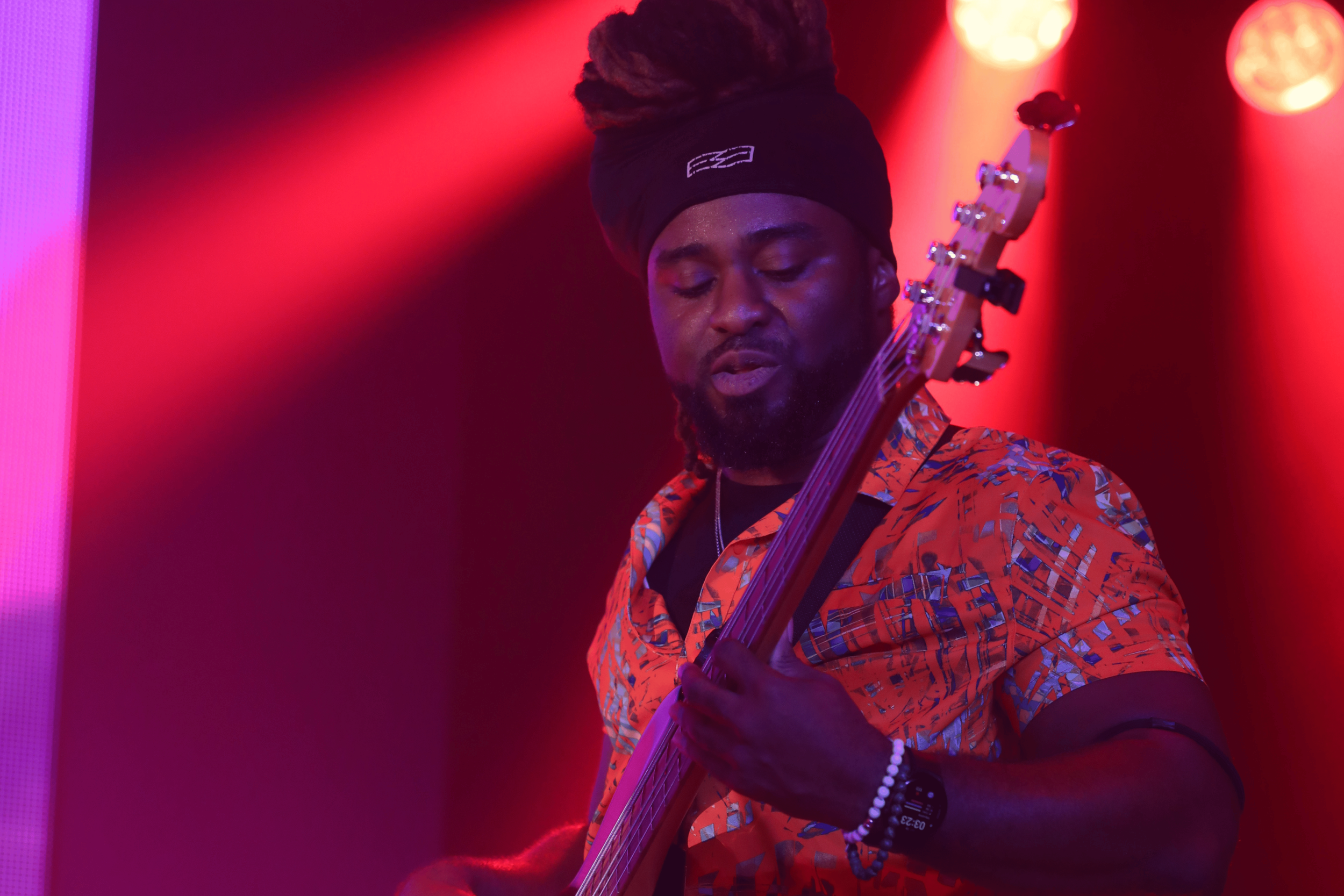 A man with dreadlocks wearing a black headband and an orange patterned shirt, playing an electric guitar on stage with red and purple stage lighting.