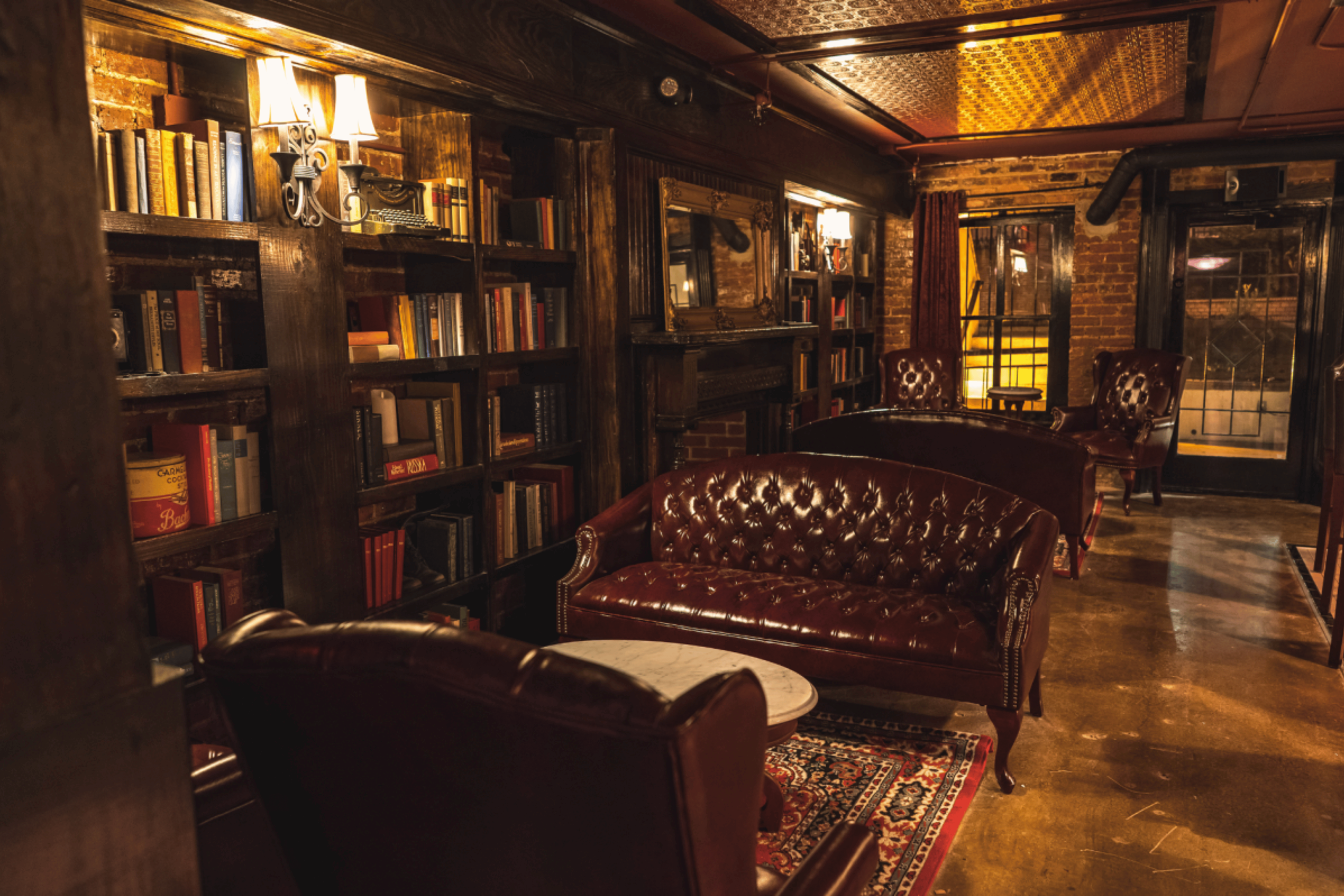 Cozy room with exposed brick walls, wooden bookshelves filled with books, leather armchairs, a fireplace, large mirror, lamps, and a rug, creating an inviting vintage ambiance.