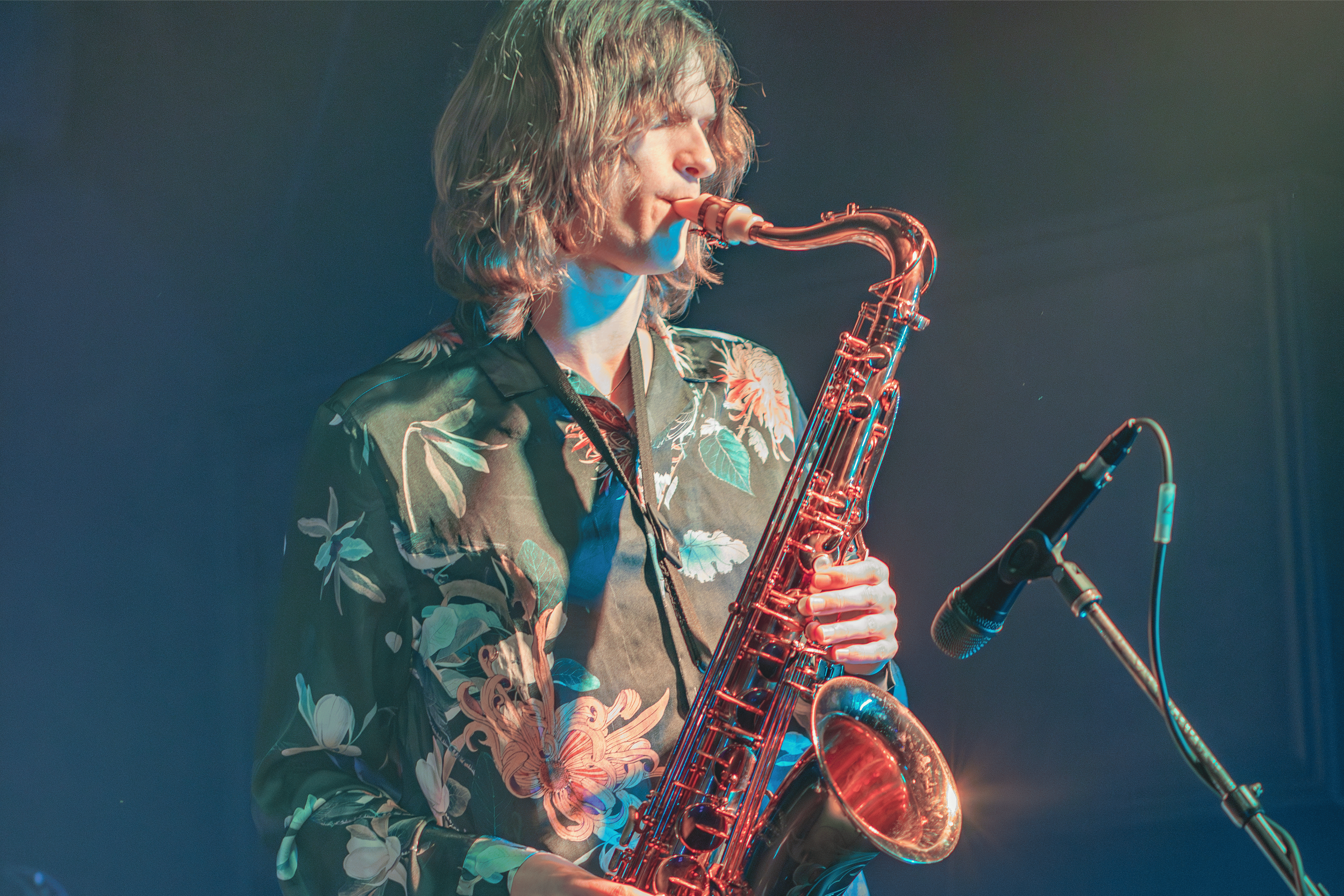 A person with shoulder-length hair playing a saxophone on stage, wearing a floral shirt, with microphones nearby and stage lighting creating a colorful atmosphere.