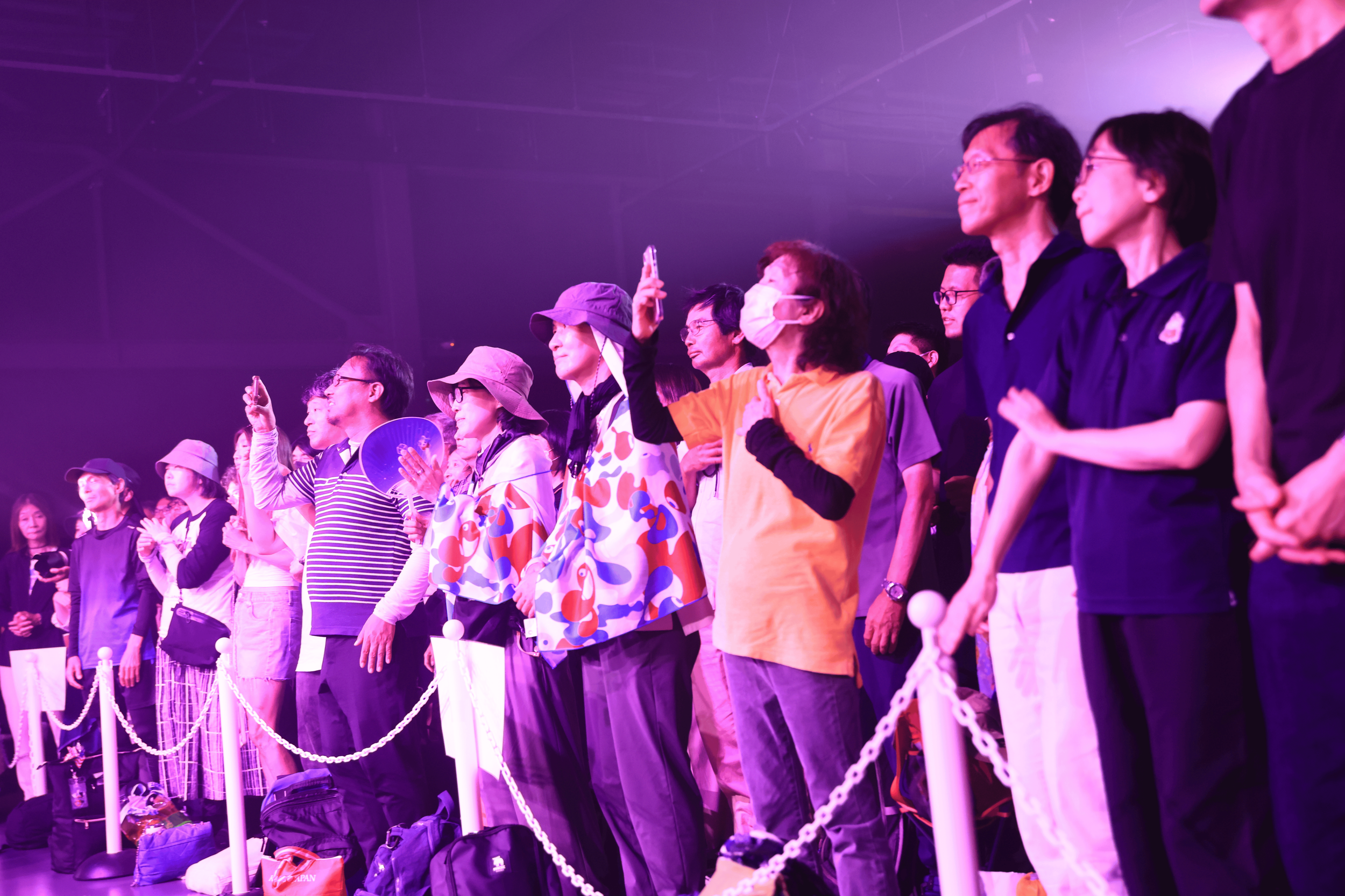Group of people standing in a row at an event, some wearing masks and holding phones, with purple lighting and a barrier rope in front of them.