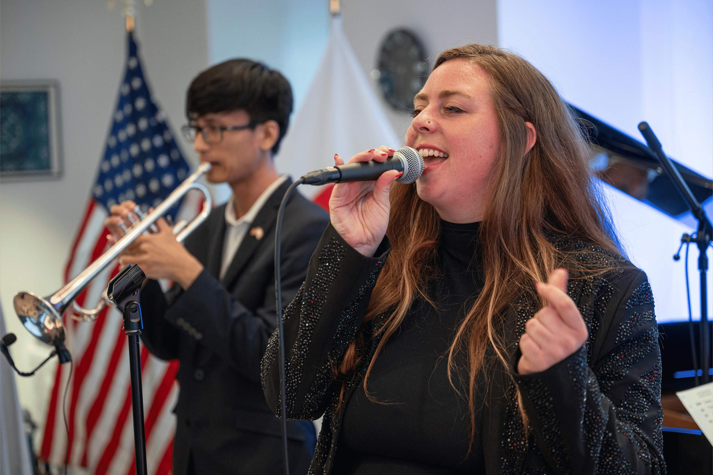 A woman singing into a microphone and a man playing a trumpet in the background. American flags are visible behind them.