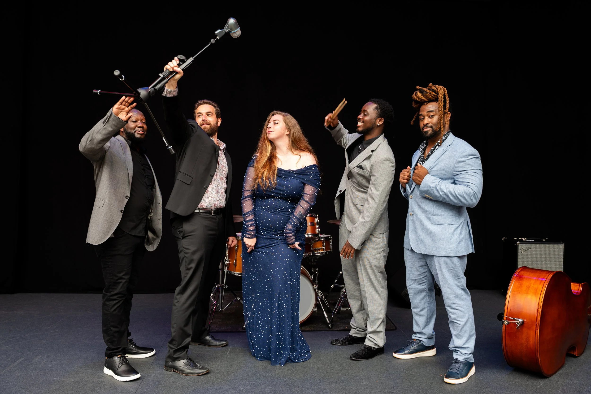 Group of five diverse musicians on stage with musical instruments, including a drum, acoustic guitar, and microphone, against a black background.