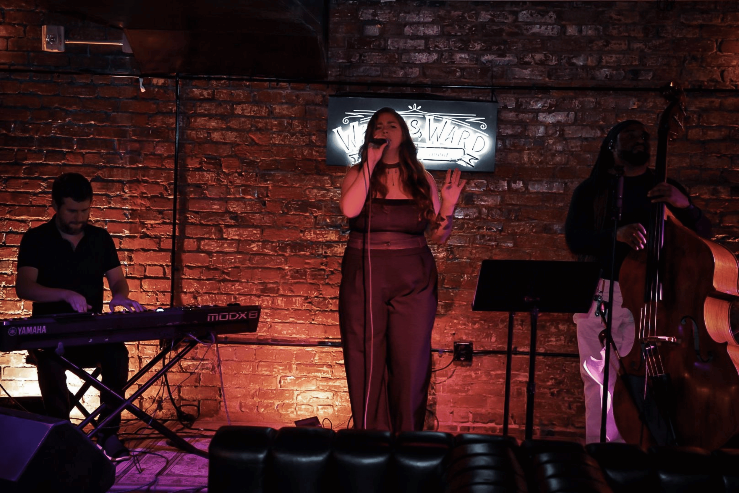 A woman singing into a microphone on a brick-walled stage with two musicians playing instruments, one on keyboard and one on double bass, in a dimly lit venue.