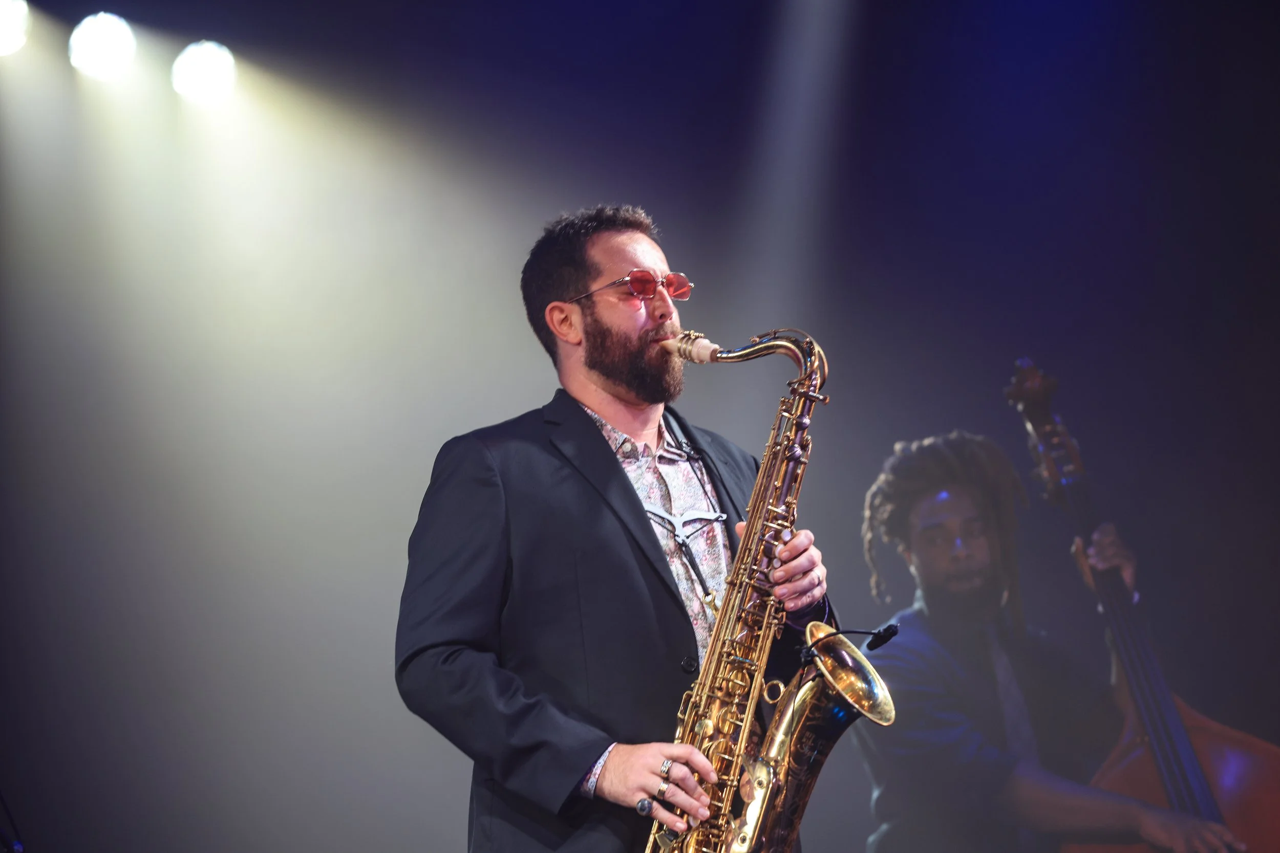 A man with dark hair, glasses, and a beard playing a gold saxophone on stage, wearing a suit jacket and floral shirt, with stage lights illuminating him in the background.
