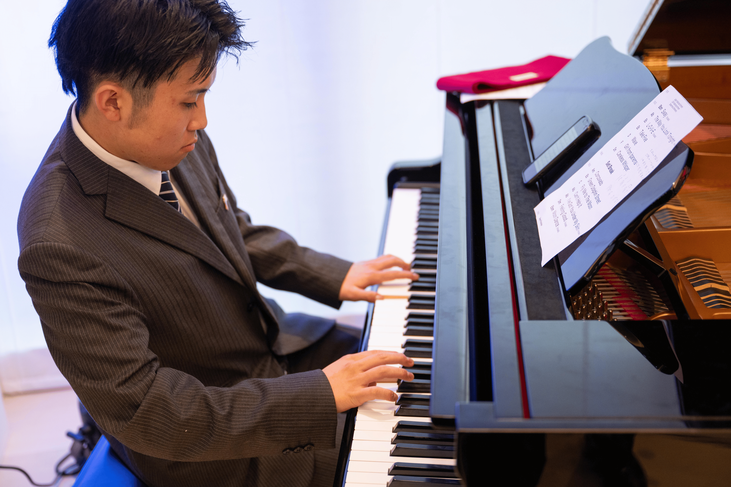 A young man in a suit playing a black piano with sheet music. The piano is situated indoors, with a pink cloth or folder on top of the piano.