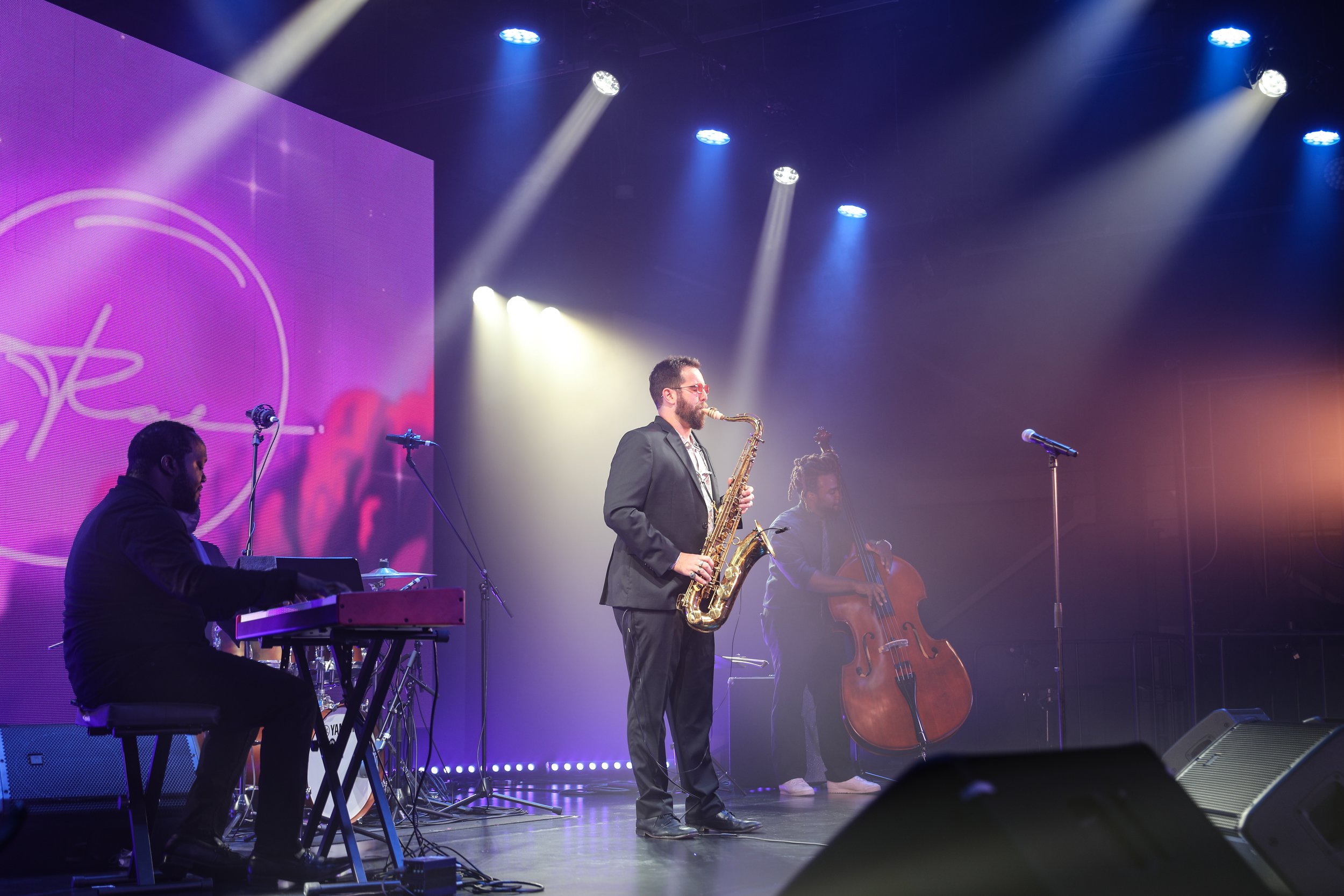 A band performing on stage with various musicians playing instruments, including a pianist, saxophonist, bassist, and double bassist, under colorful stage lighting.