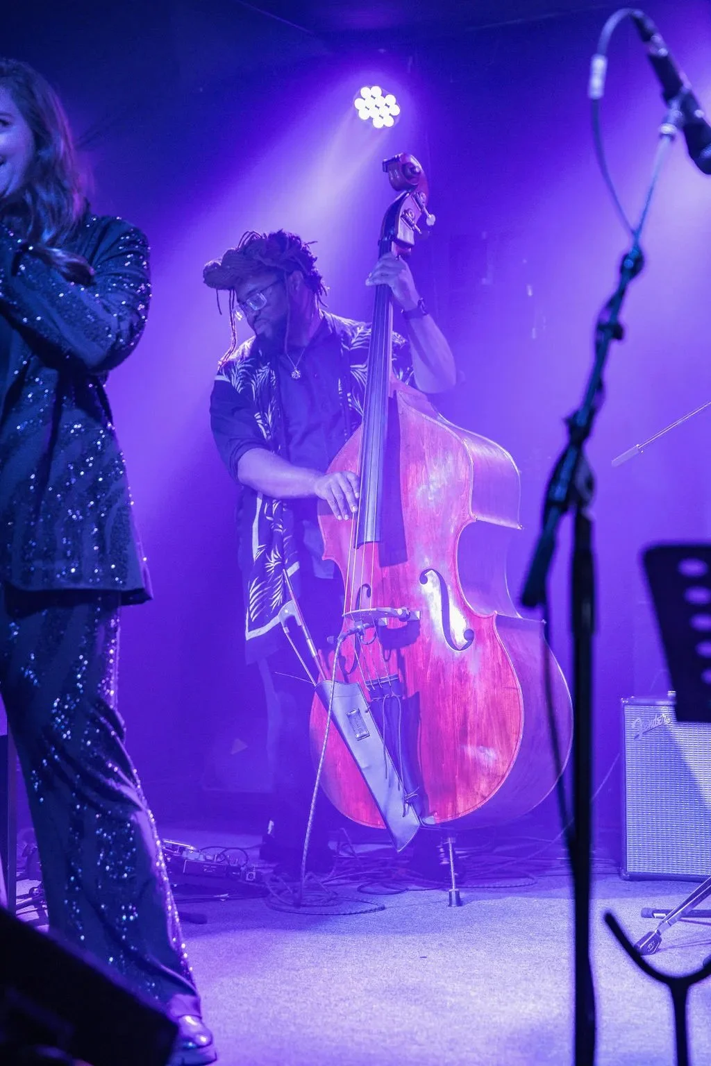 Musician playing double bass on stage under purple and blue lighting, wearing glasses and a patterned shirt.