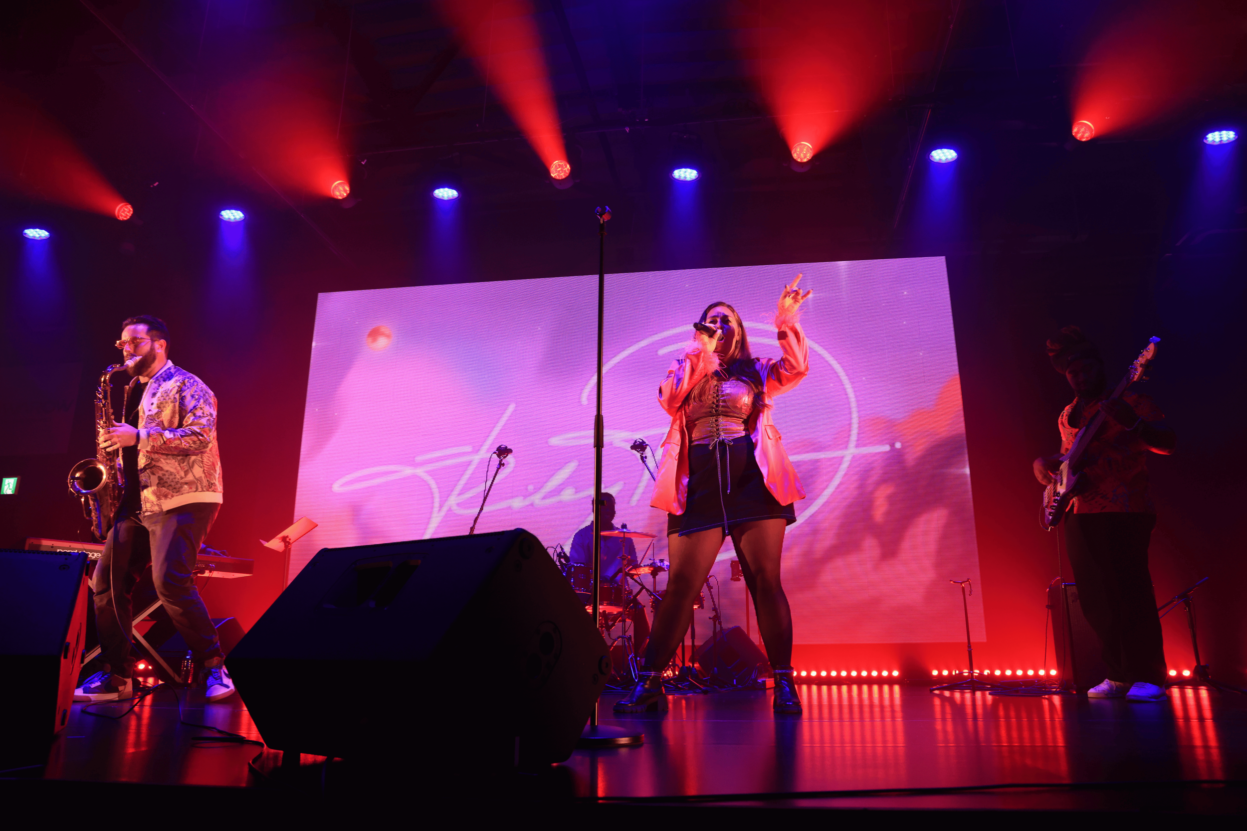 Performer singing on stage with a microphone, accompanied by musicians, under colorful red and blue stage lights, with a large digital screen displaying graphics behind them.