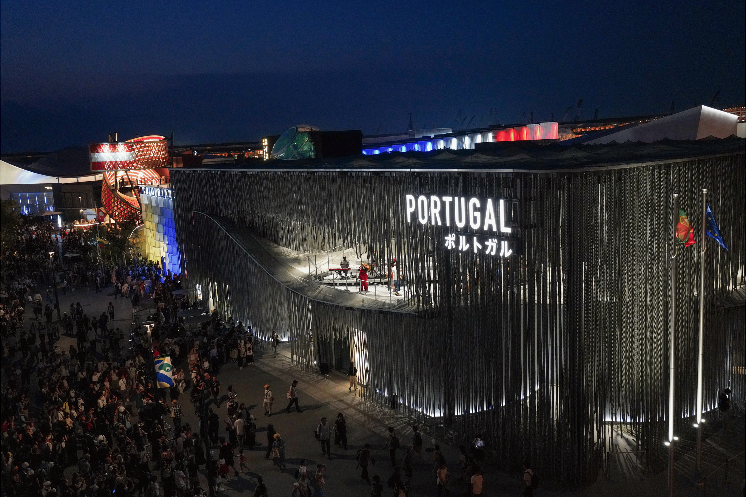 Night view of a modern building or exhibition center with illuminated sign that says 'PORTUGAL' and flags, including the Portuguese flag, outside. The scene shows a large crowd of people gathered around the building.