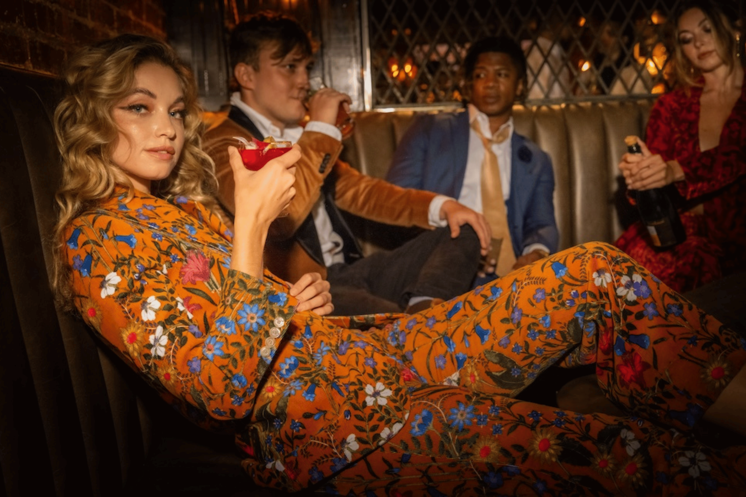 A woman in an orange floral dress with blonde hair is sitting at a bar, holding a cocktail, while three men in suits sit nearby and converse, in what appears to be a dimly-lit lounge or bar setting.