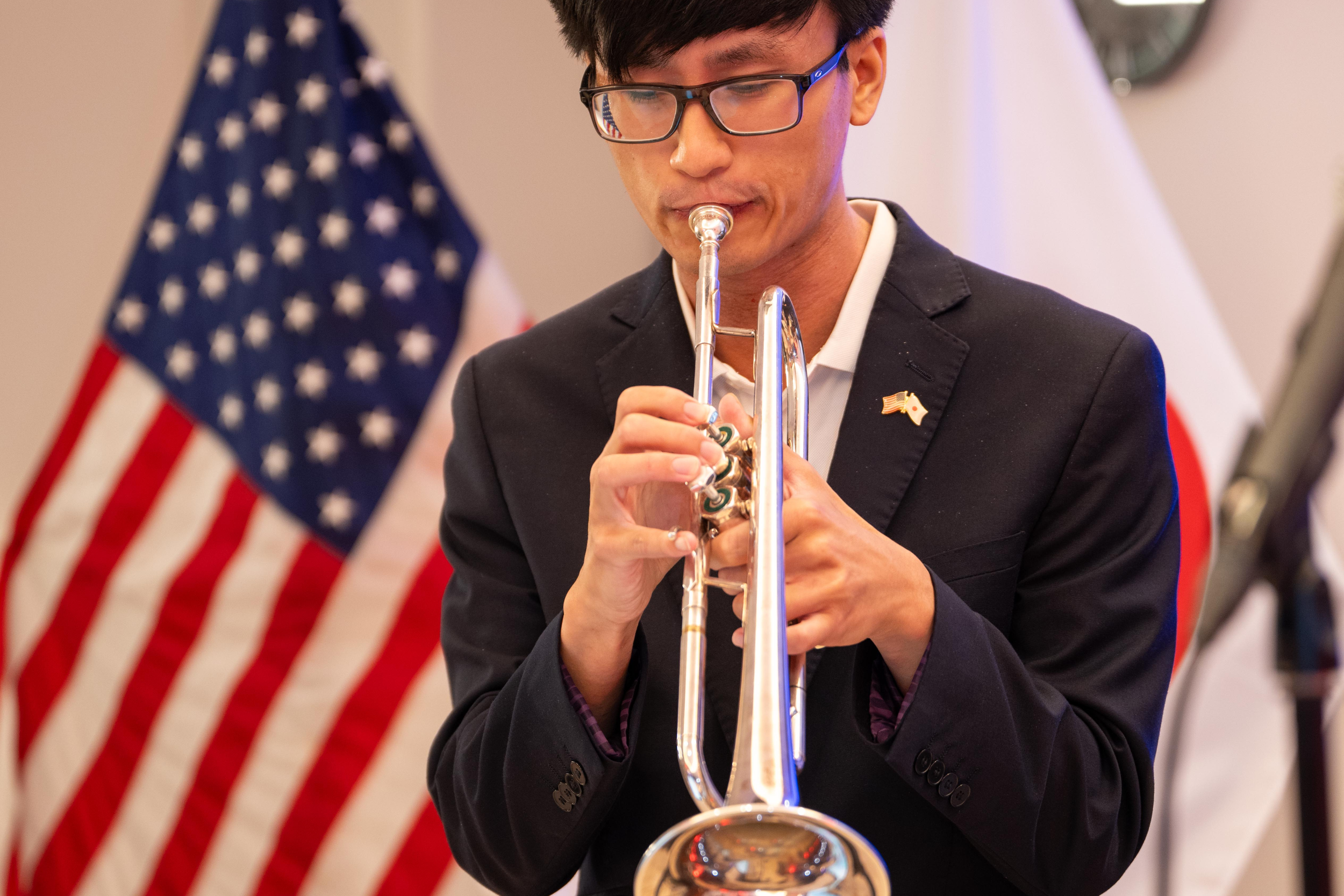 A young man with glasses playing the trumpet in front of an American flag at an indoor event.