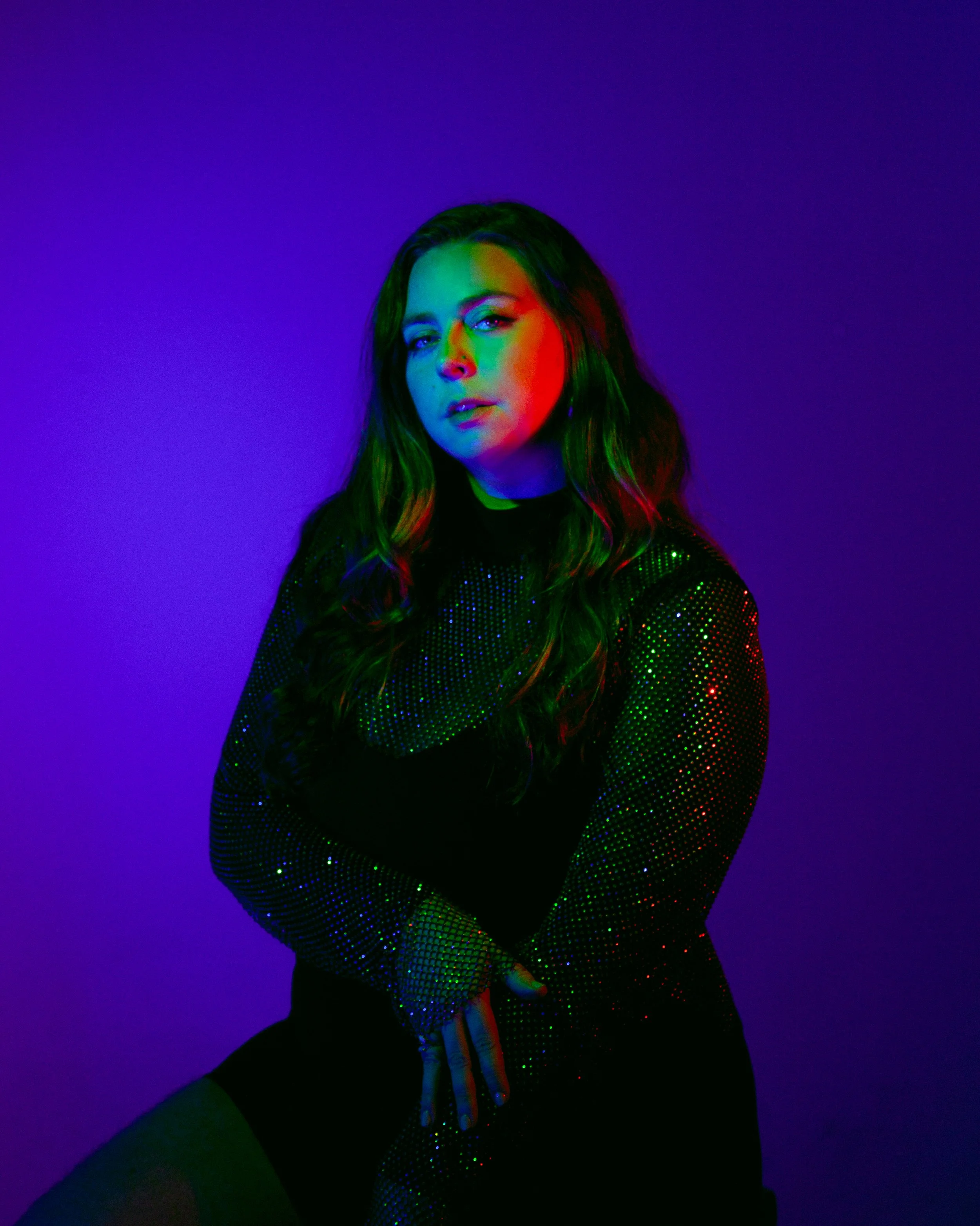 A woman with long wavy hair stands in front of a purple background, illuminated by multicolored lights, wearing a black and silver sparkly top.