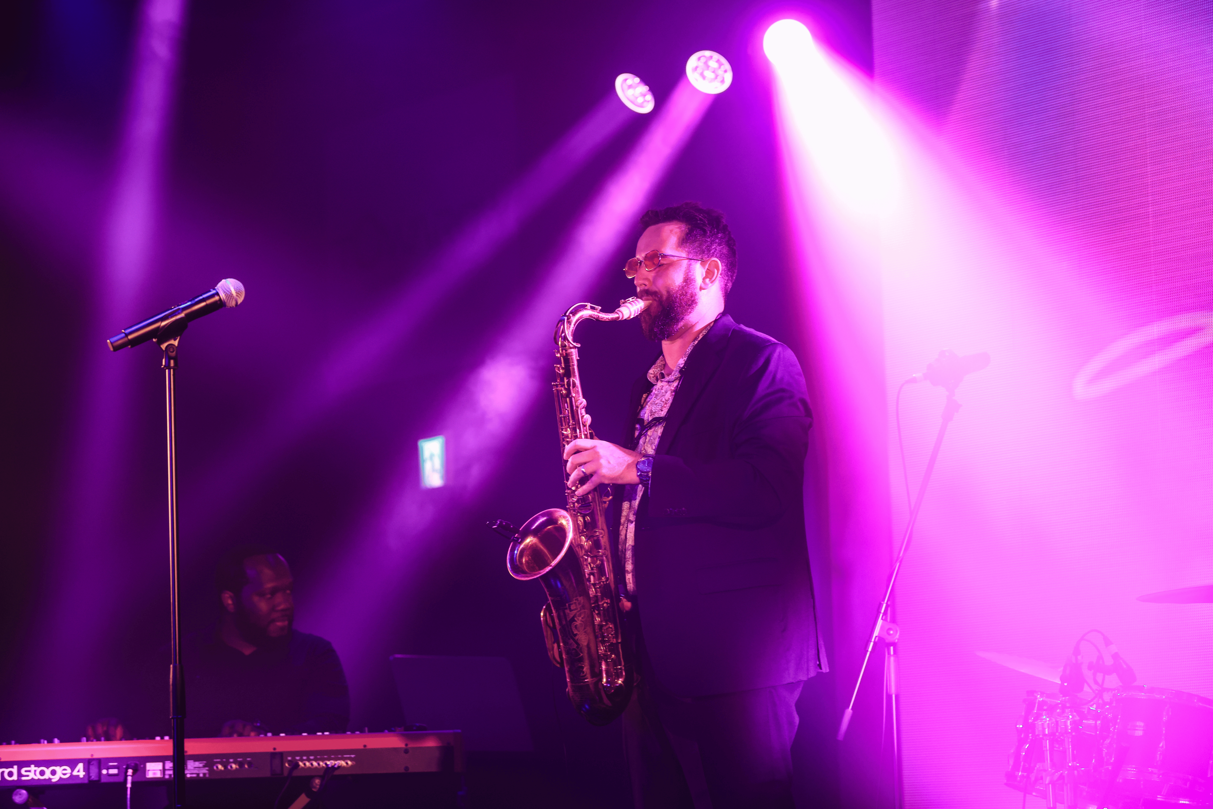 A man in glasses and a suit playing the saxophone on stage with purple and pink stage lighting.