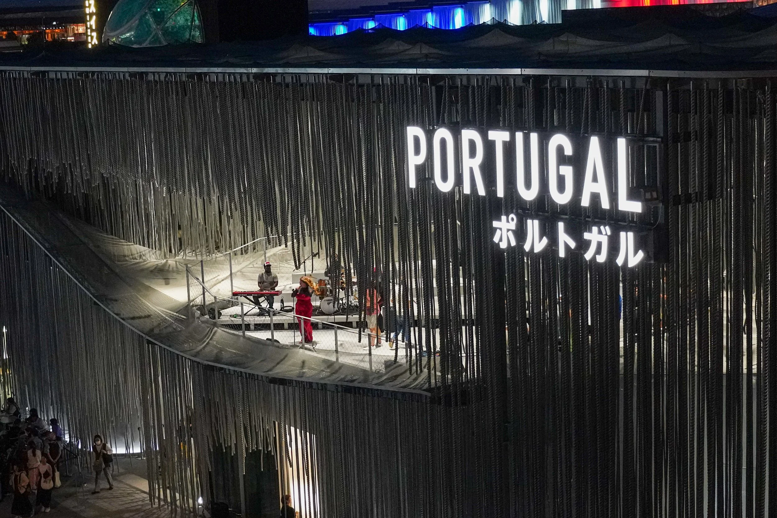 Night scene of a modern building with a large illuminated sign that says 'PORTUGAL' in English and Japanese characters. There are people walking at the entrance, and others are on a platform with band members performing.