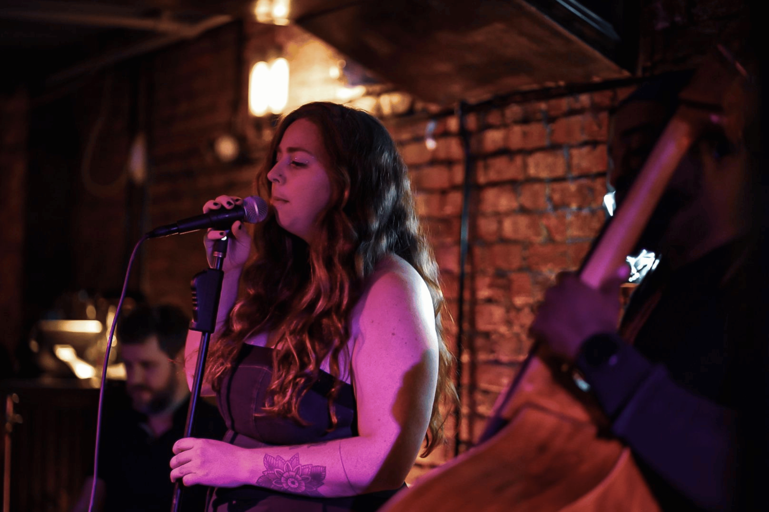 A woman with long curly hair singing into a microphone at a dimly lit venue with brick walls.