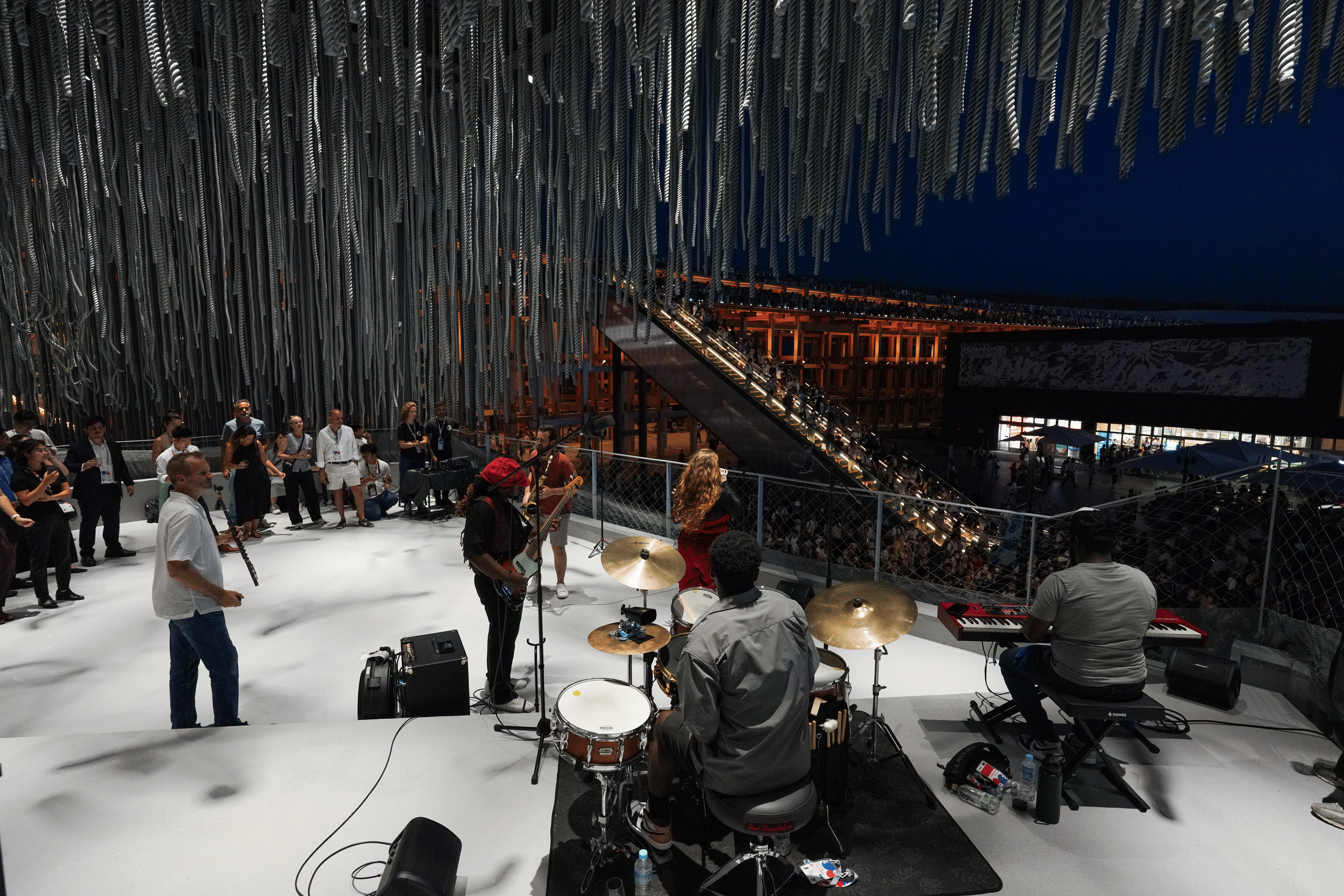 A musical performance inside a modern building with tall, decorative ceiling elements. The stage has several musicians playing instruments, including drums, guitar, and keyboard. People are standing and observing the performance, and the background s