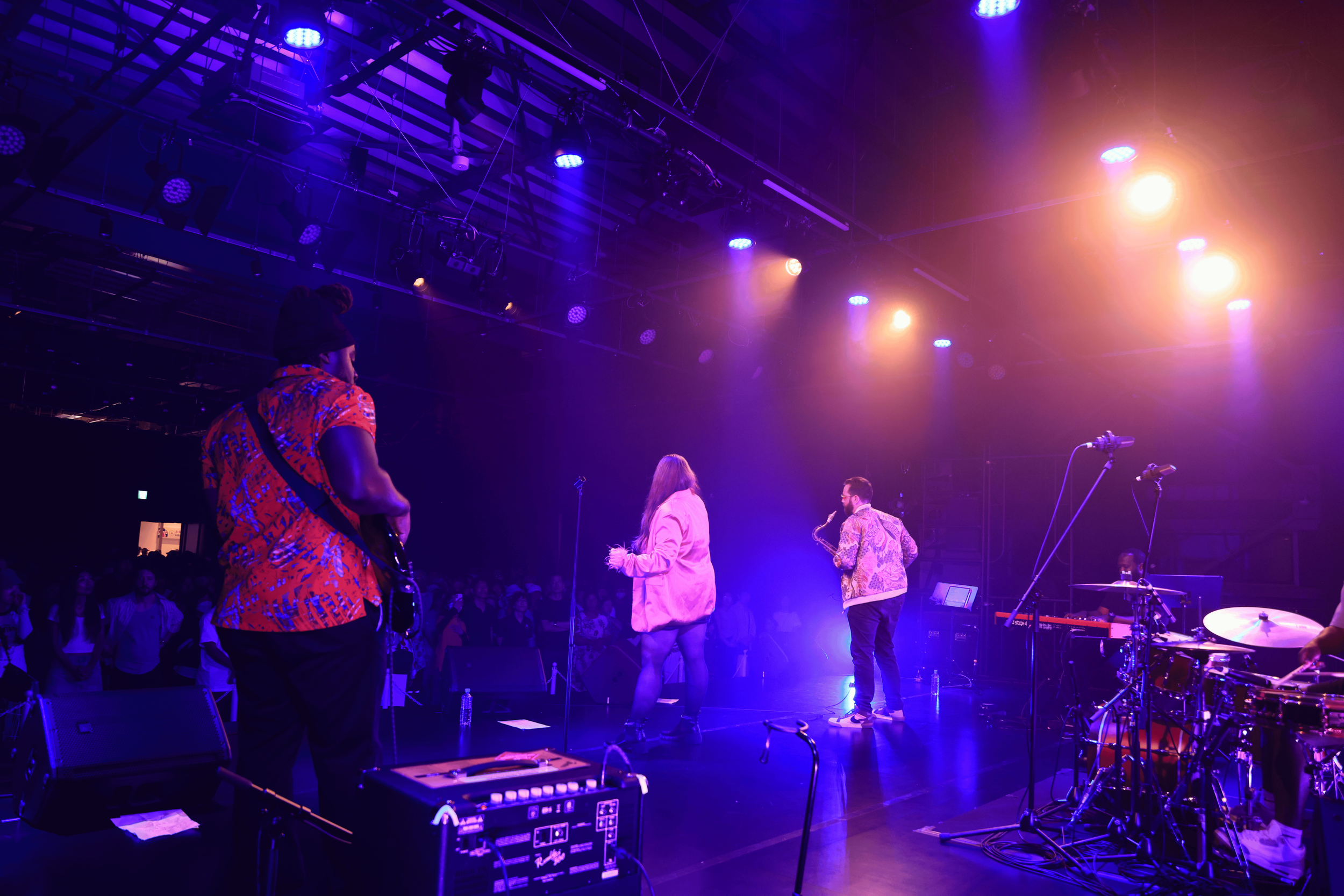 Musicians performing on a stage with colorful purple and orange lighting. Audience watching in the background with a drum set and microphones visible.