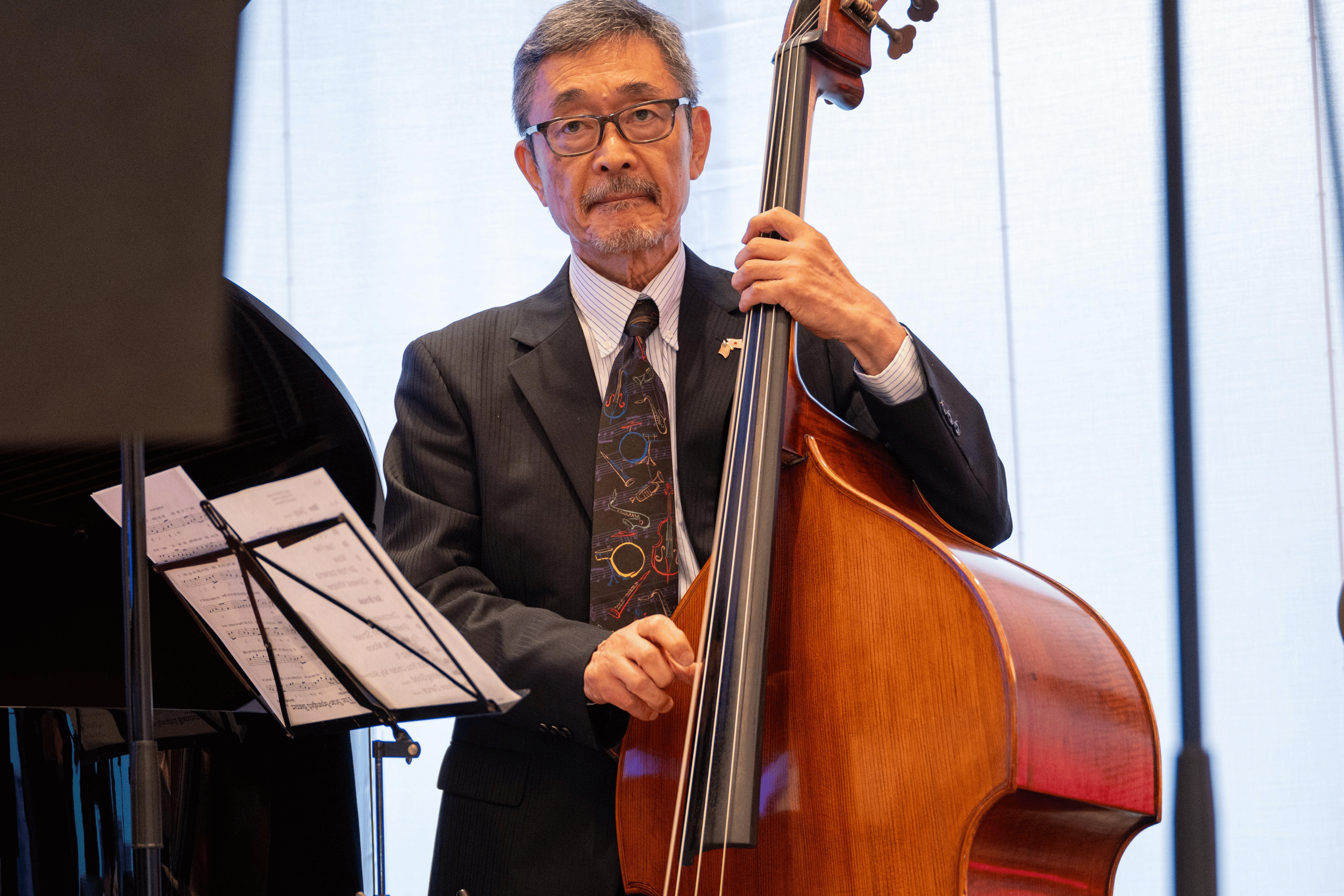 An older man wearing glasses and a suit playing a double bass in a room with large windows.