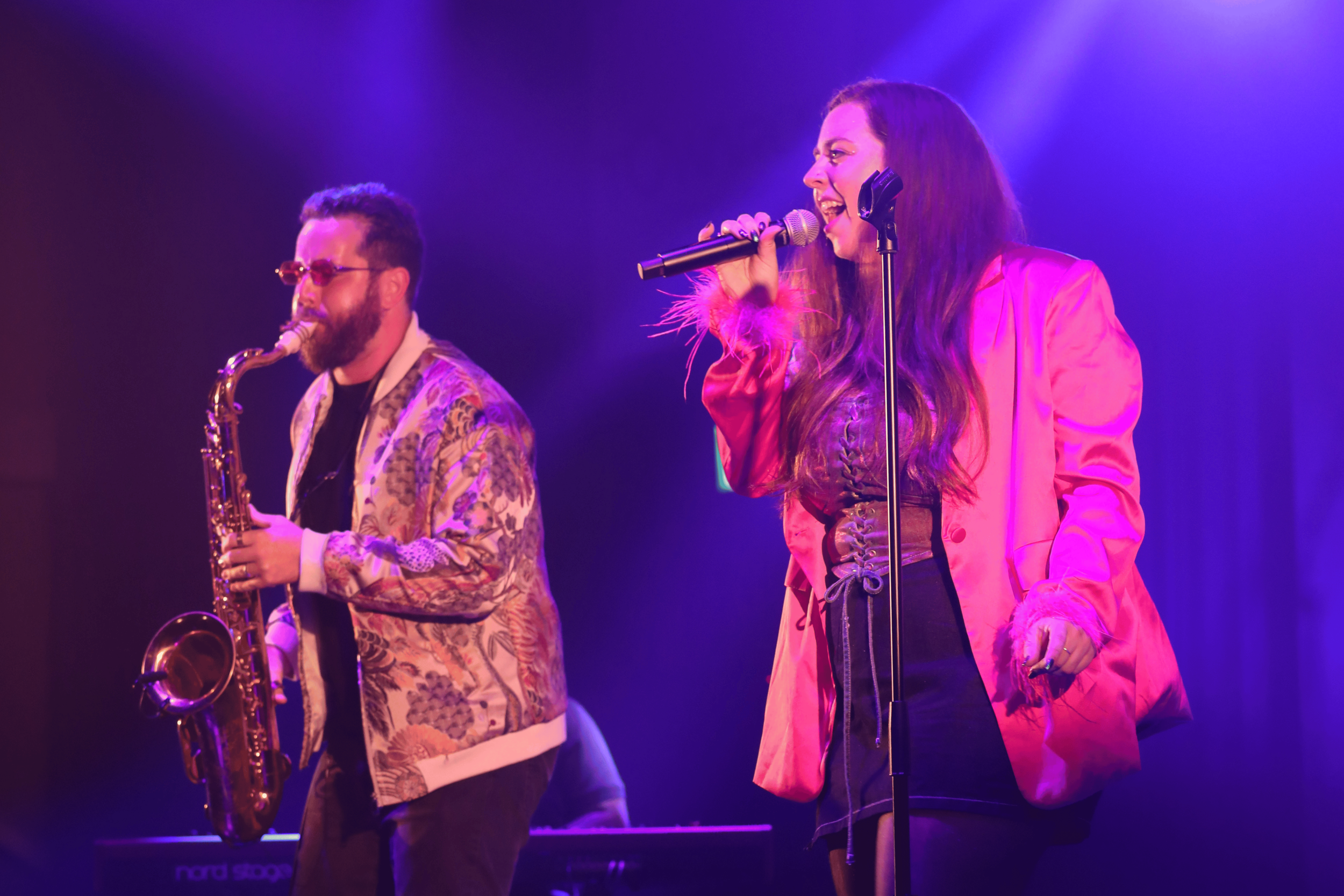A woman singing into a microphone on stage with purple and blue lighting, wearing a pink jacket with fur cuffs, and a man playing a saxophone nearby, both enjoying the performance.
