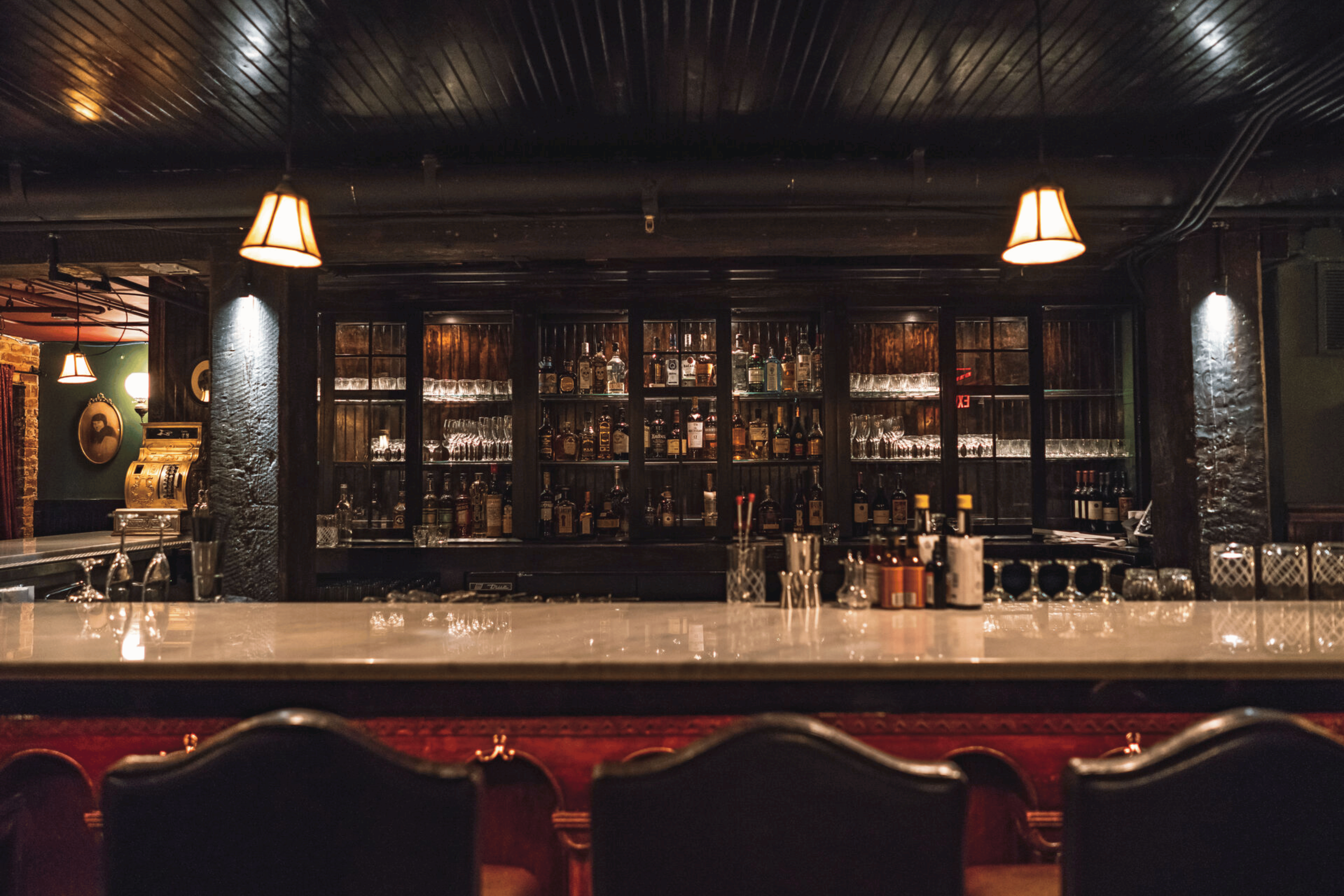 A dimly lit bar with a dark wooden counter and a back wall filled with various bottles of alcohol and glasses. The ceiling has a dark wooden finish with hanging lamps providing warm lighting.