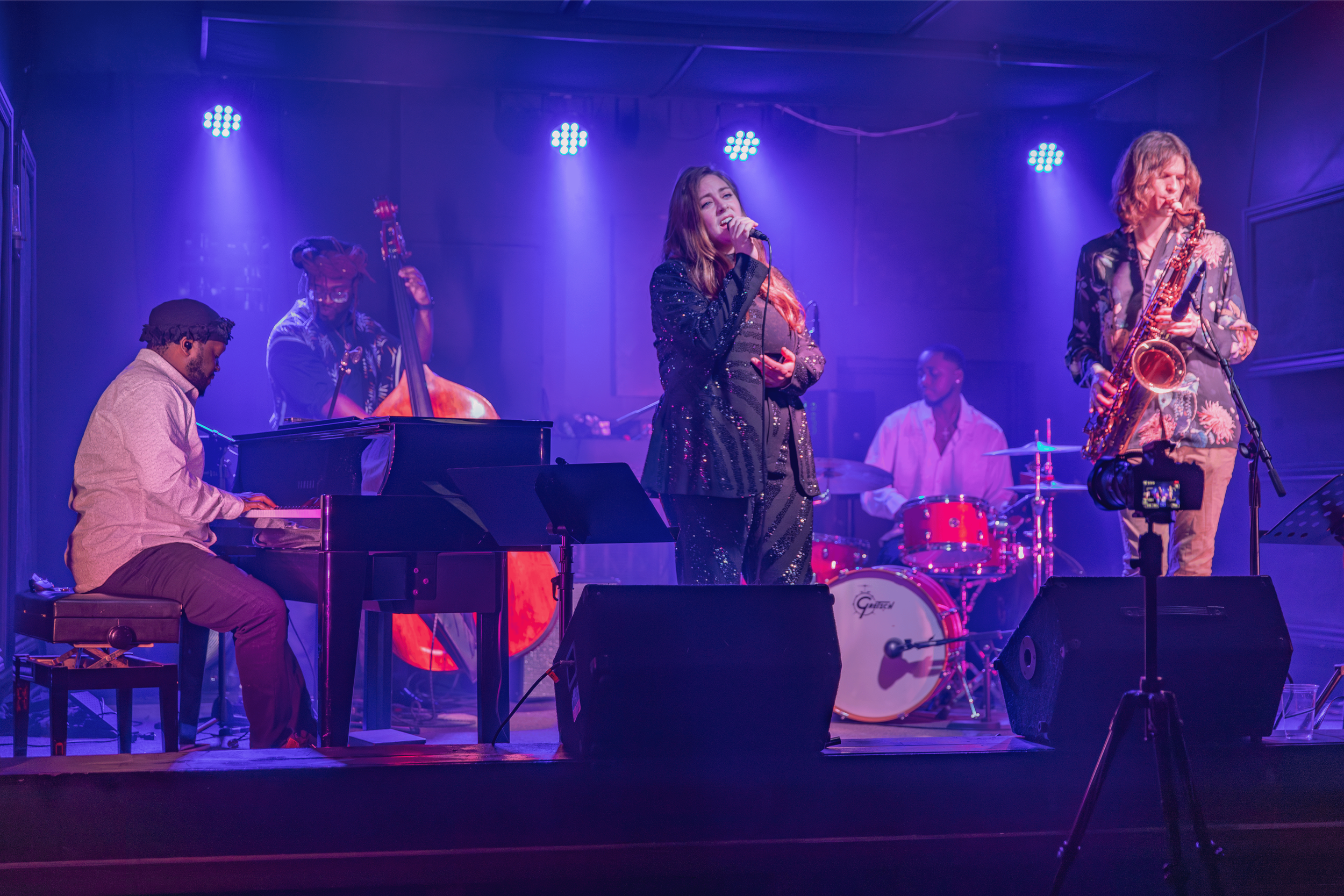 A musical band performing on stage under blue and purple lighting. The band includes a pianist, a double bass player, a singer, a drummer, and a saxophonist.