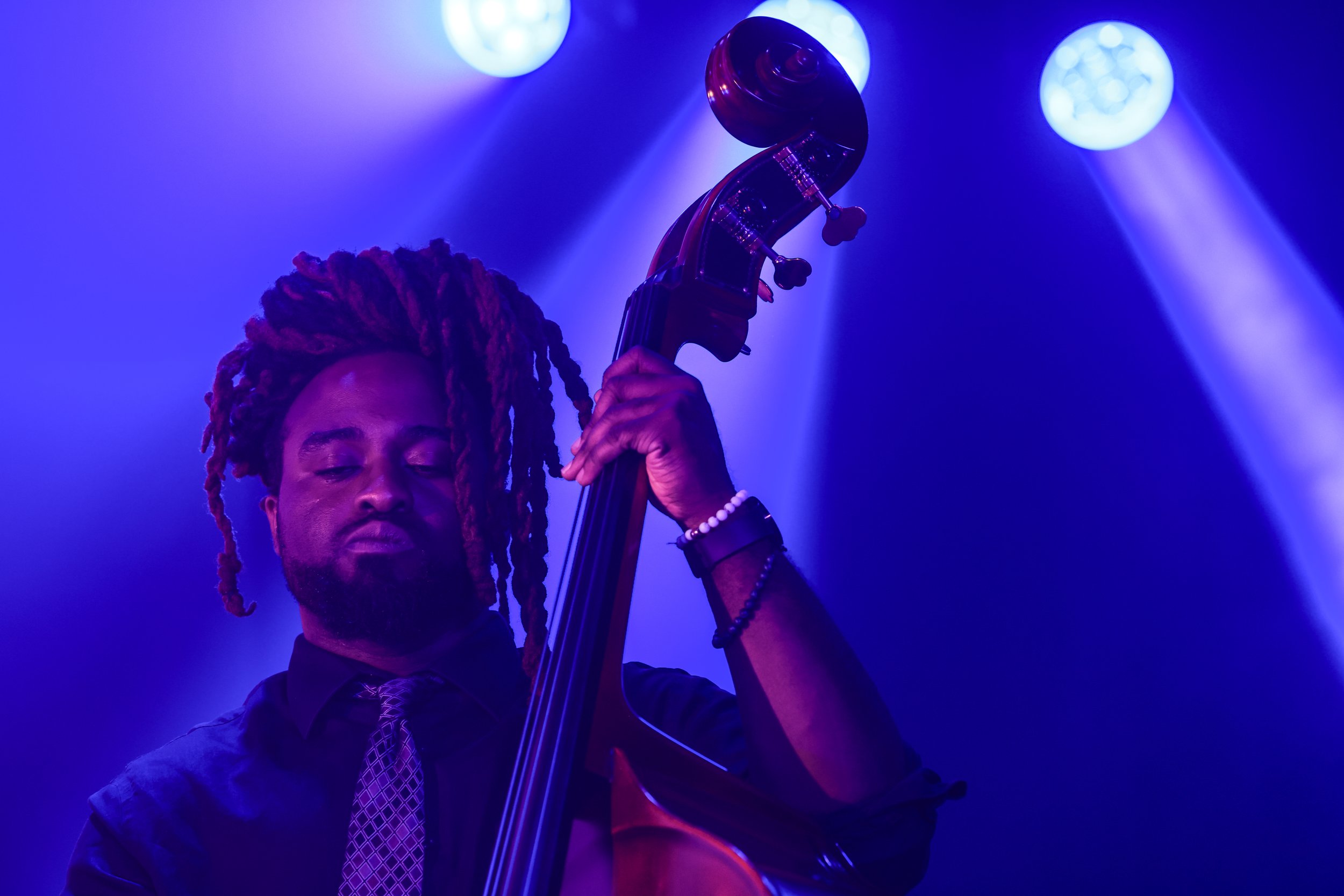 A person with closed eyes playing a double bass under purple and blue stage lights.